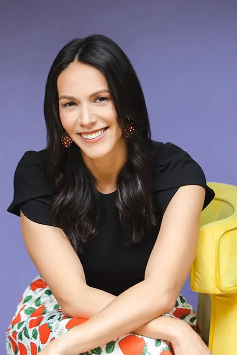 A woman with long black hair, smiling, wearing a black top and floral skirt, sitting next to a yellow chair against a purple background.