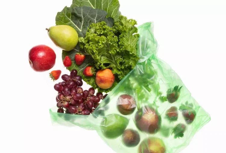 Fresh fruits and vegetables on a white background, with some in a green plastic bag. Items include grapes, strawberries, apples, lettuce, cabbage, a pear, peaches, and onions.