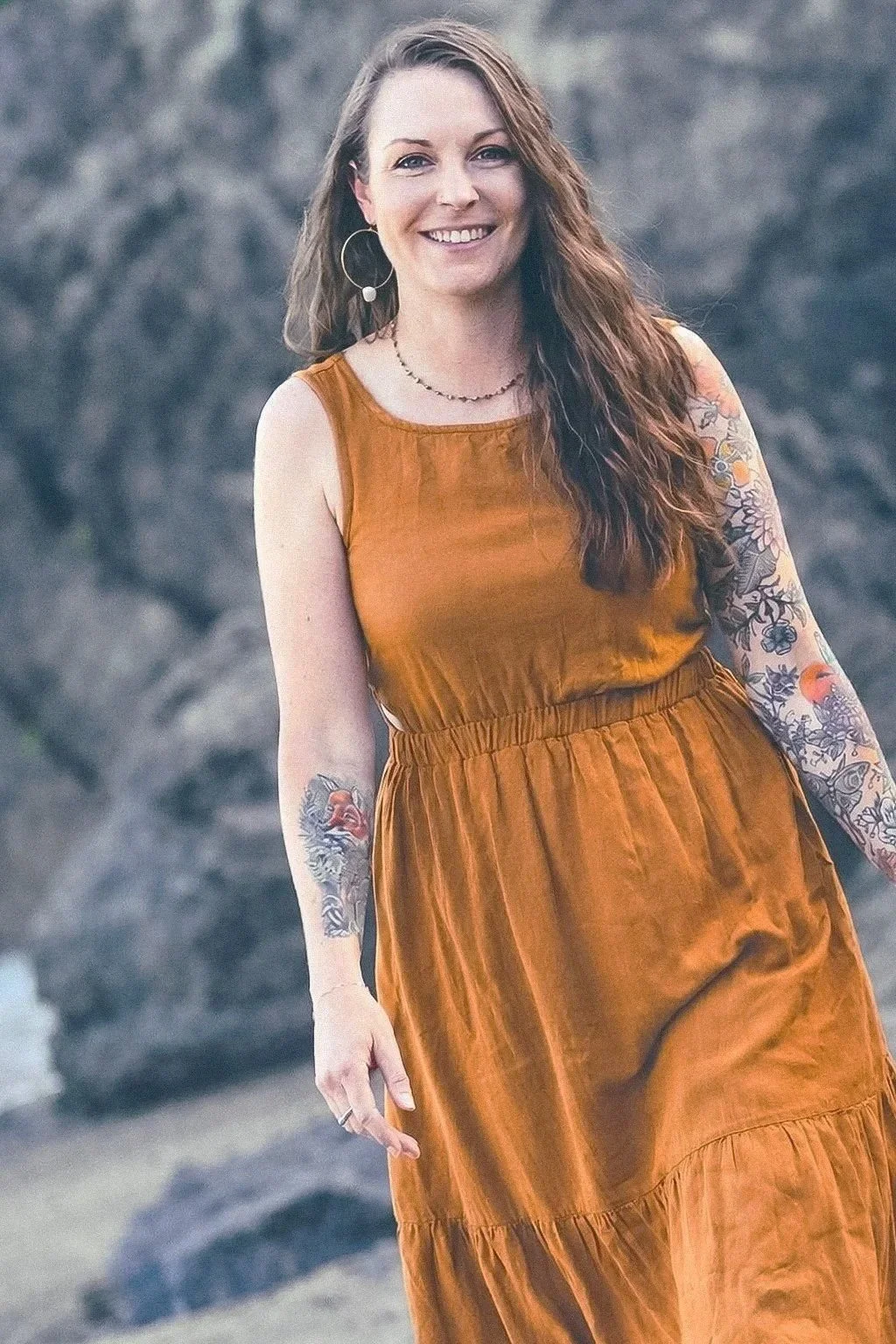 A woman with long, wavy brown hair, wearing a sleeveless brown dress with a gathered waist, standing outdoors with a rocky background. She is smiling and has tattoos on both arms and hoop earrings.