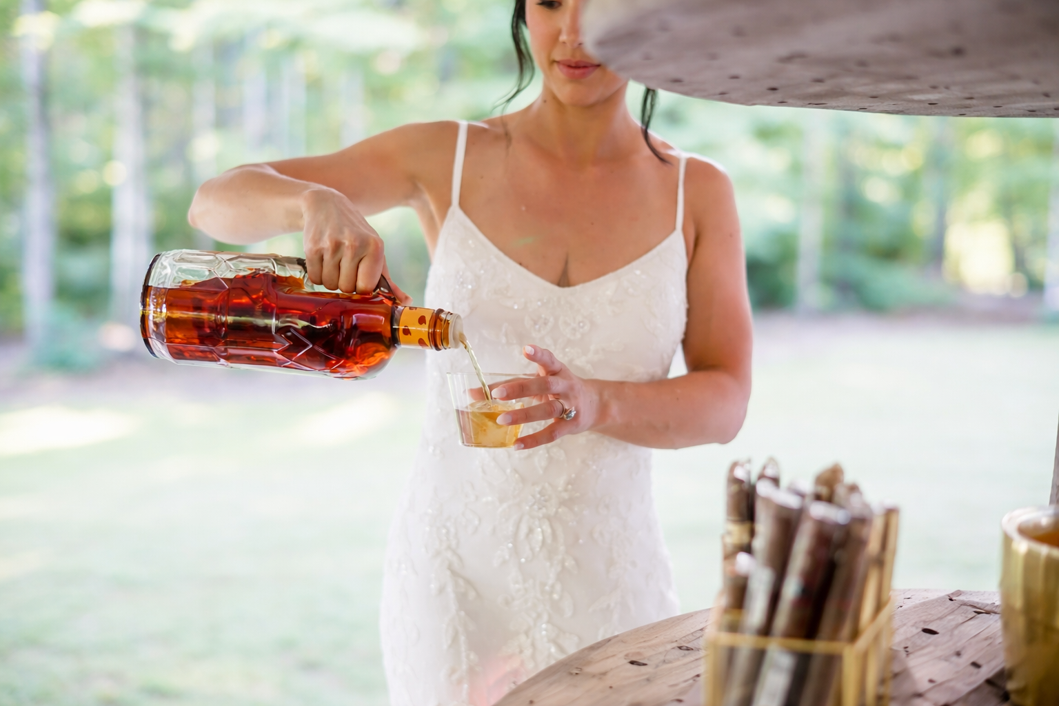 A woman in a white dress pours a drink from a bottle into a glass outdoors in a wooded area.