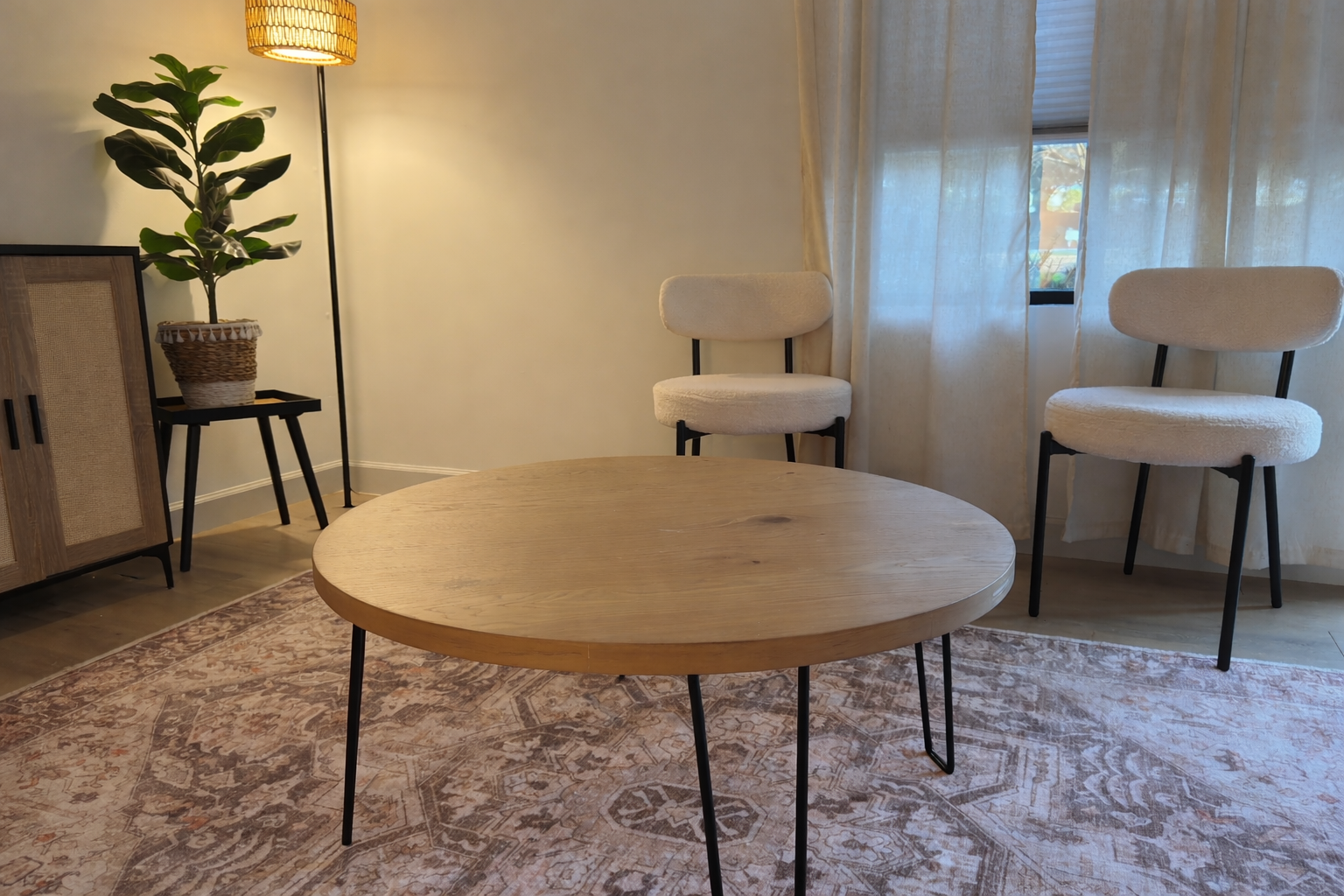 Living room with a round wooden coffee table, two beige upholstered chairs, a wooden cabinet, a potted plant on a small black table, a tall floor lamp, curtains, and a window with partially open blinds.