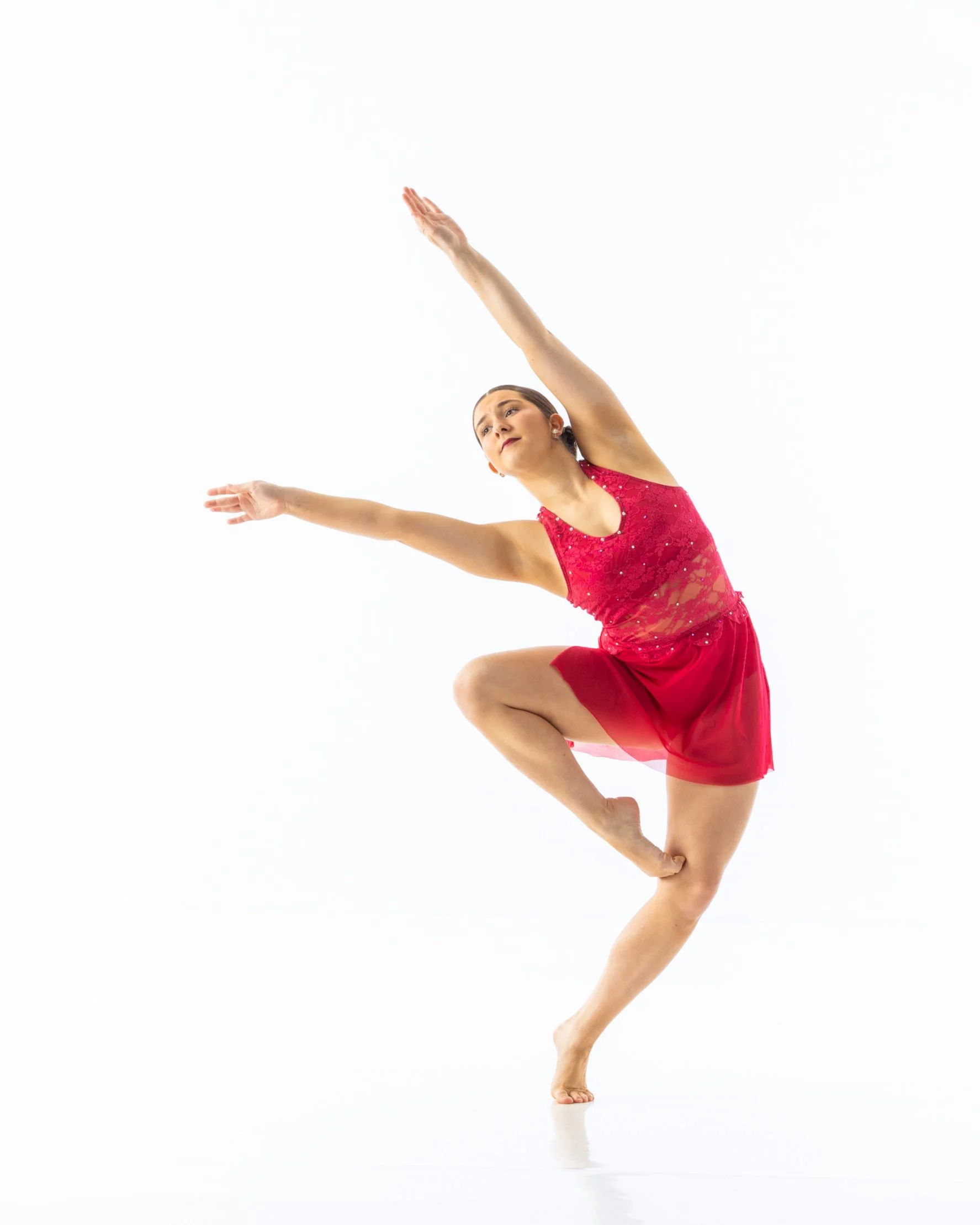A female figure skater in a red dress performs a move with one leg bent and her arm extended upward against a plain white background.