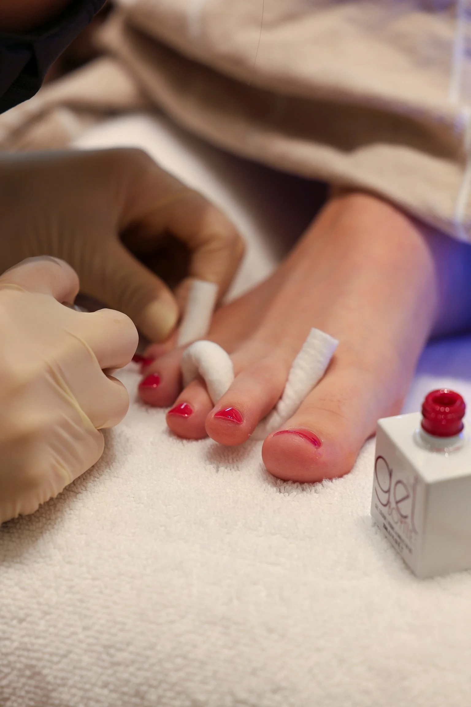 Close-up of a person having their nails painted in a salon, with a nail technician applying red nail polish to their fingernails.