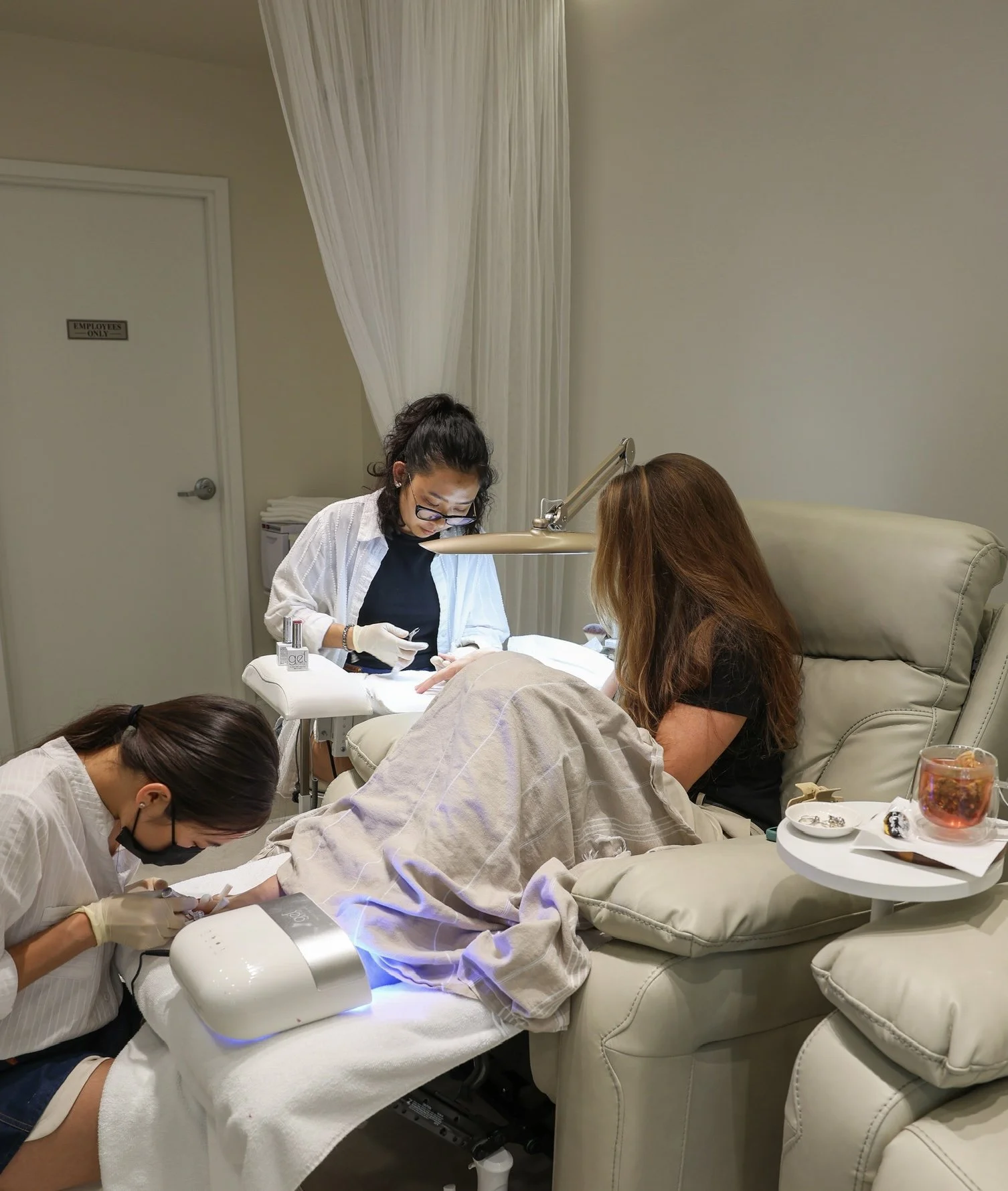 A group of women in a medical or spa setting getting hand or nail treatments while lying on reclined chairs. One woman is receiving care from a technician, with others working nearby.