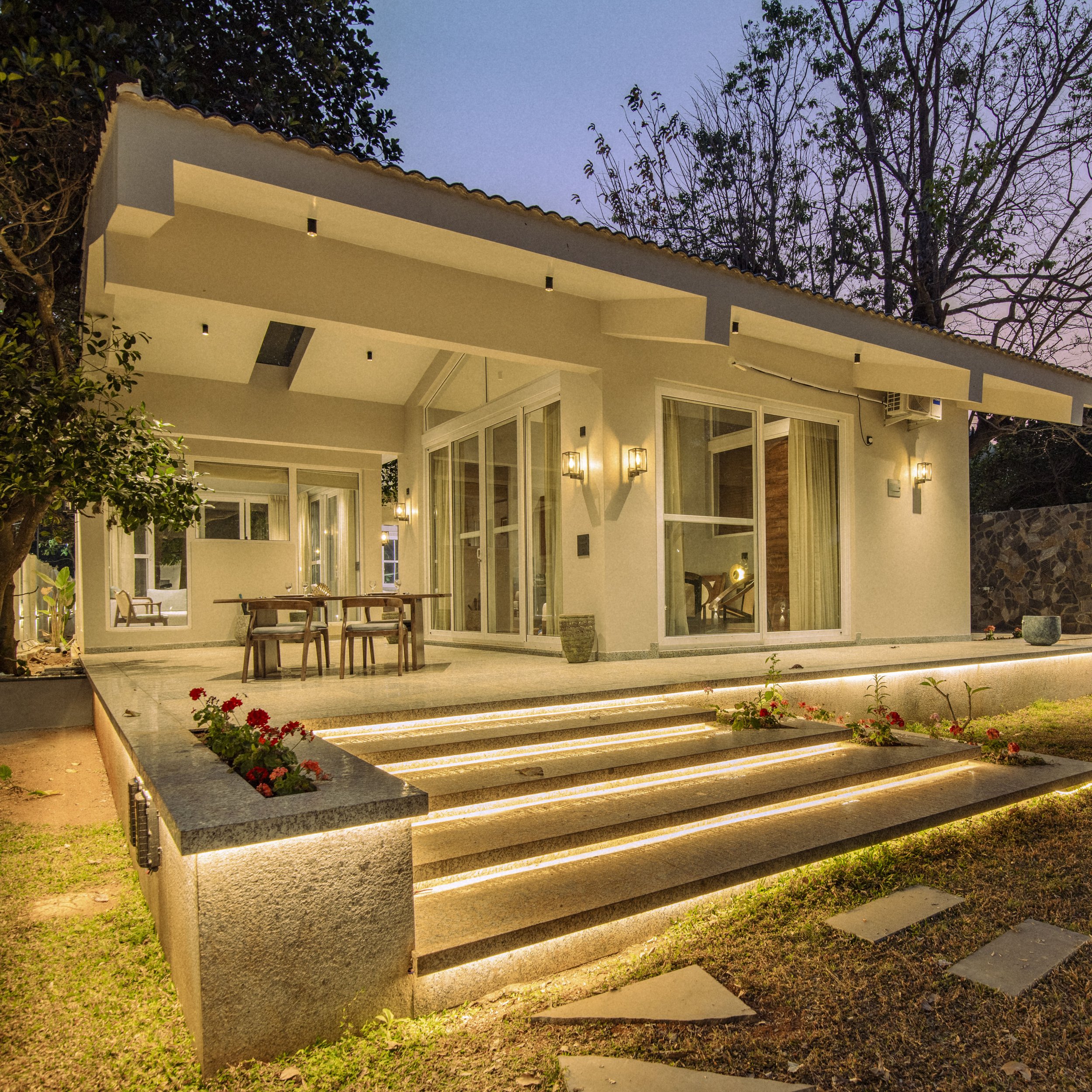 A modern house exterior at dusk with stairs lit by LED strip lights, patio furniture, plants, and large glass doors and windows.