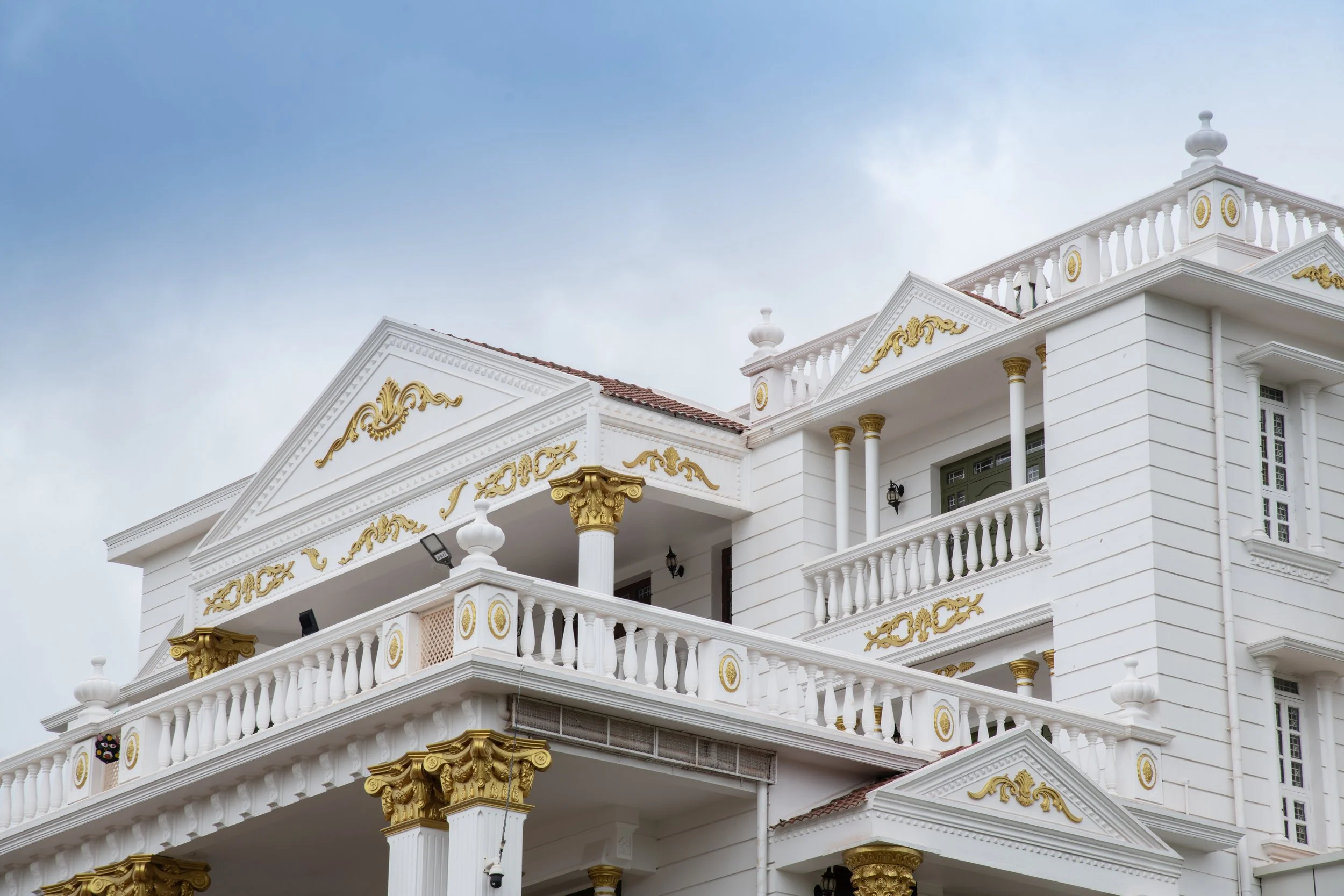 A white, ornate multi-story mansion with gold accents on architectural details, columns, and balustrades, set against a background of a cloudy sky.
