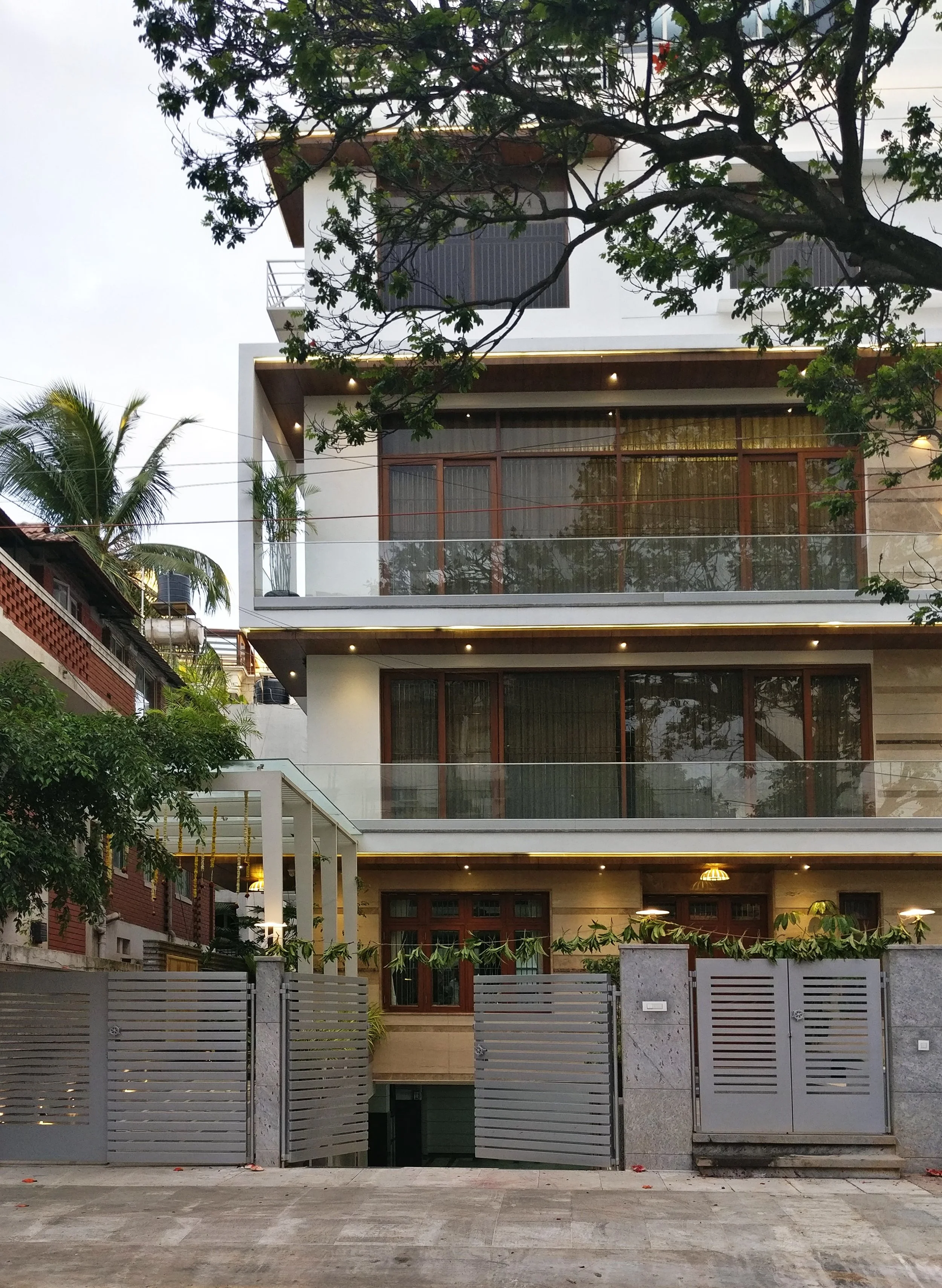 Modern multi-story residential building with glass balconies, large windows, wooden accents, and a gated entrance, surrounded by trees.