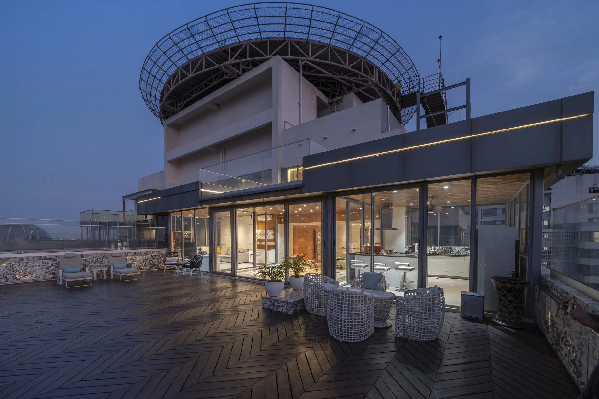 Rooftop terrace of a modern building with outdoor seating, potted plants, and glass walls showcasing the interior kitchen and living space, under a twilight sky.