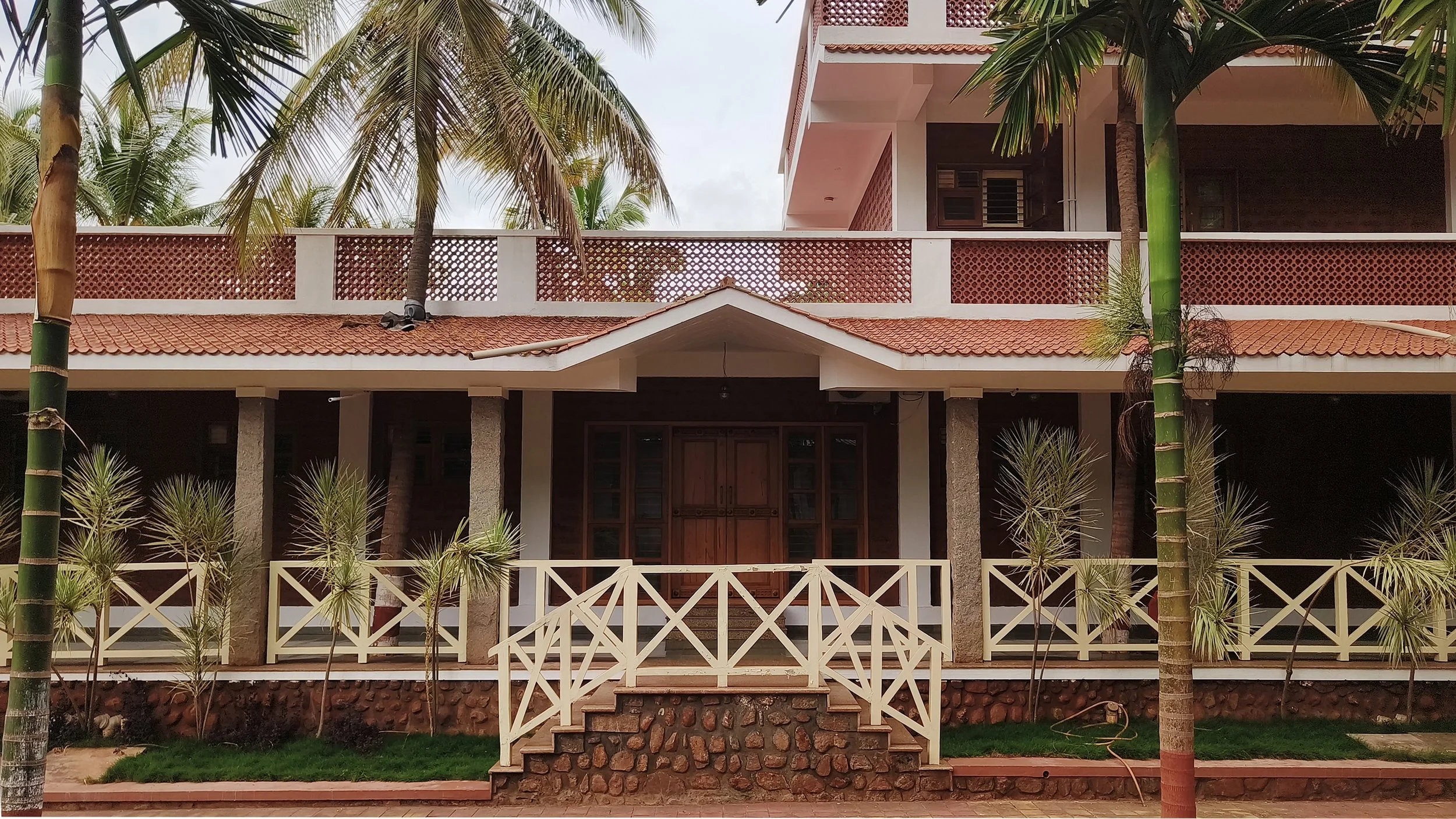 Front view of a modern two-story house with a red-tiled roof, surrounded by palm trees, and a white porch railing with steps leading to the entrance.