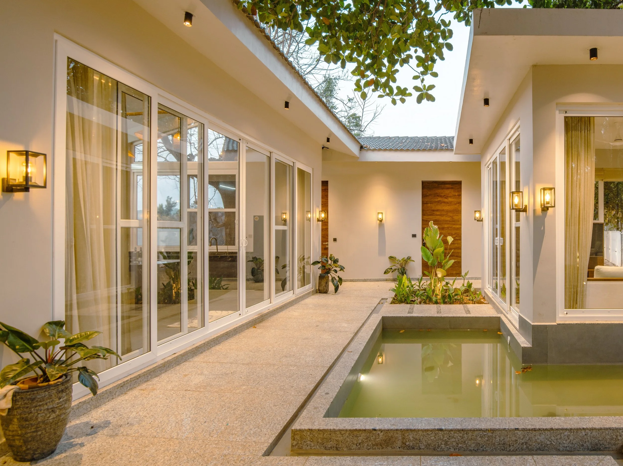 Modern indoor-outdoor living space with large glass doors, a small reflective pool, potted plants, and warm wall-mounted lighting.