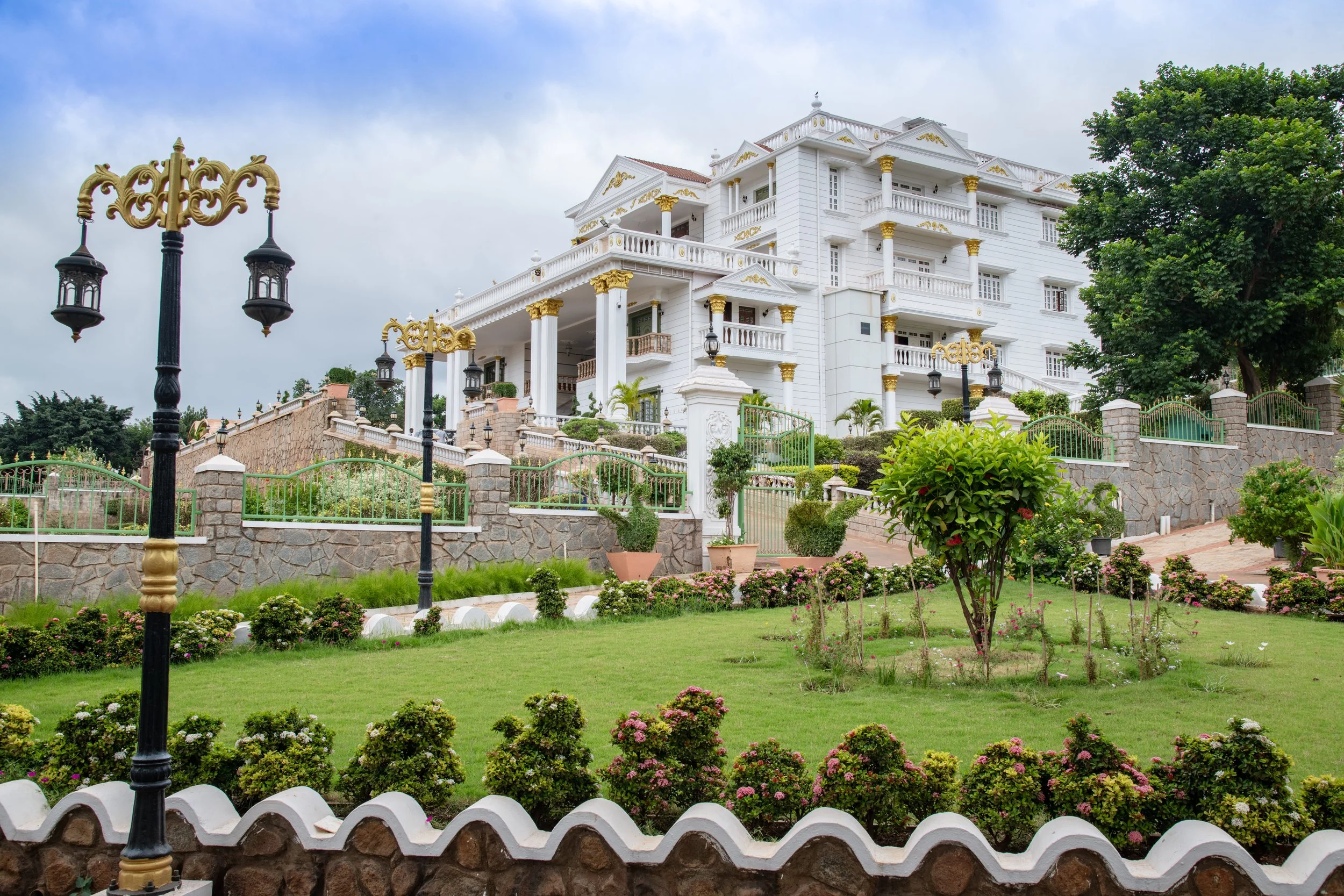 Large white mansion with gold accents, surrounded by a landscaped garden with trees, shrubs, and flowers, and decorative black and gold lamp posts.