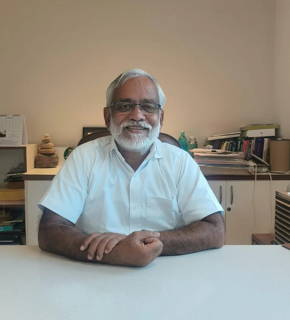 A smiling older man with gray hair and beard, wearing glasses and a white shirt, sitting at a desk in an office with books and supplies in the background.