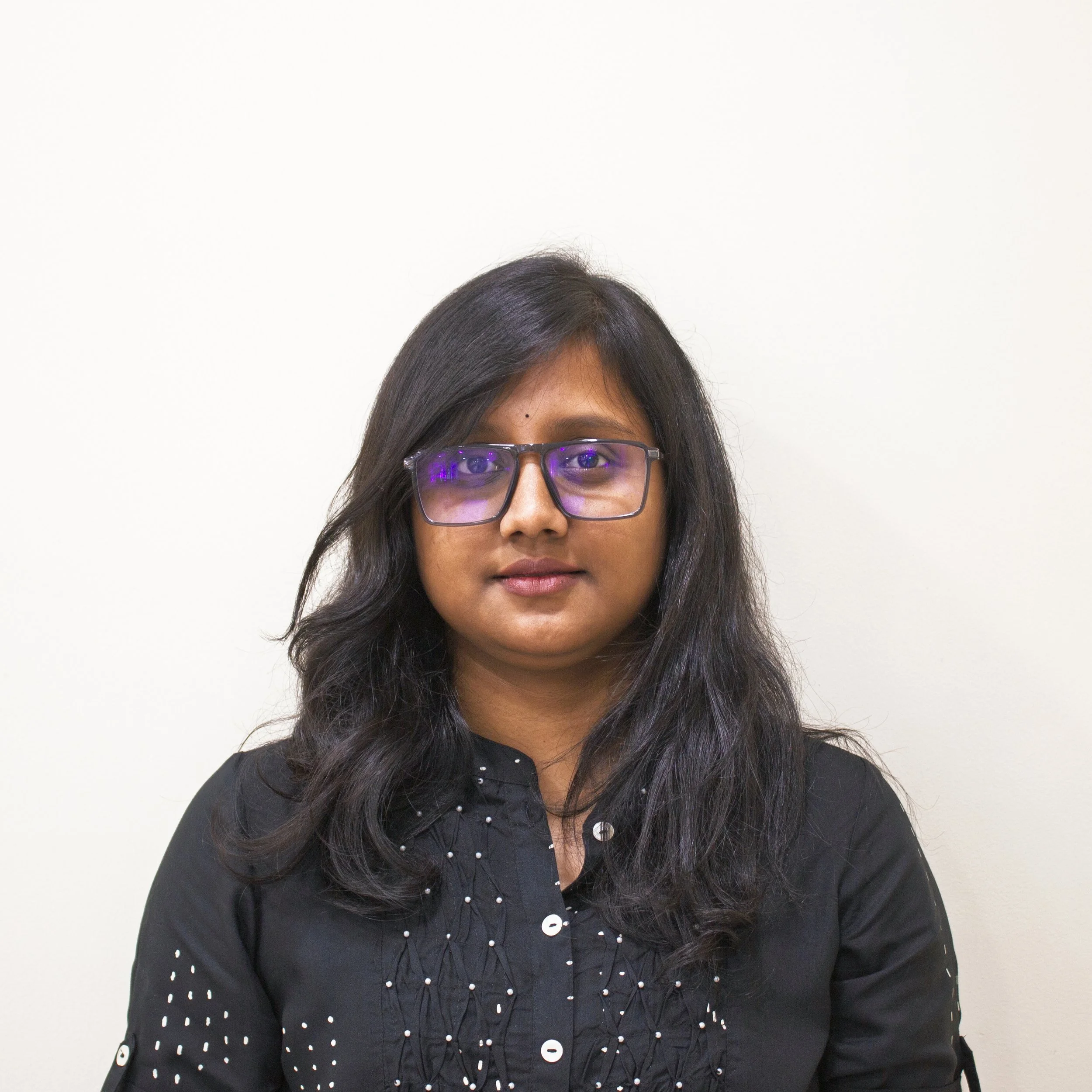 A woman with short blonde hair, wearing a sleeveless rust-colored top and beige pants, sitting on a beige cushioned bench writing in a notebook. There is a large plant on her left, abstract artwork on her right, and sunlight casting shadows on the wall.