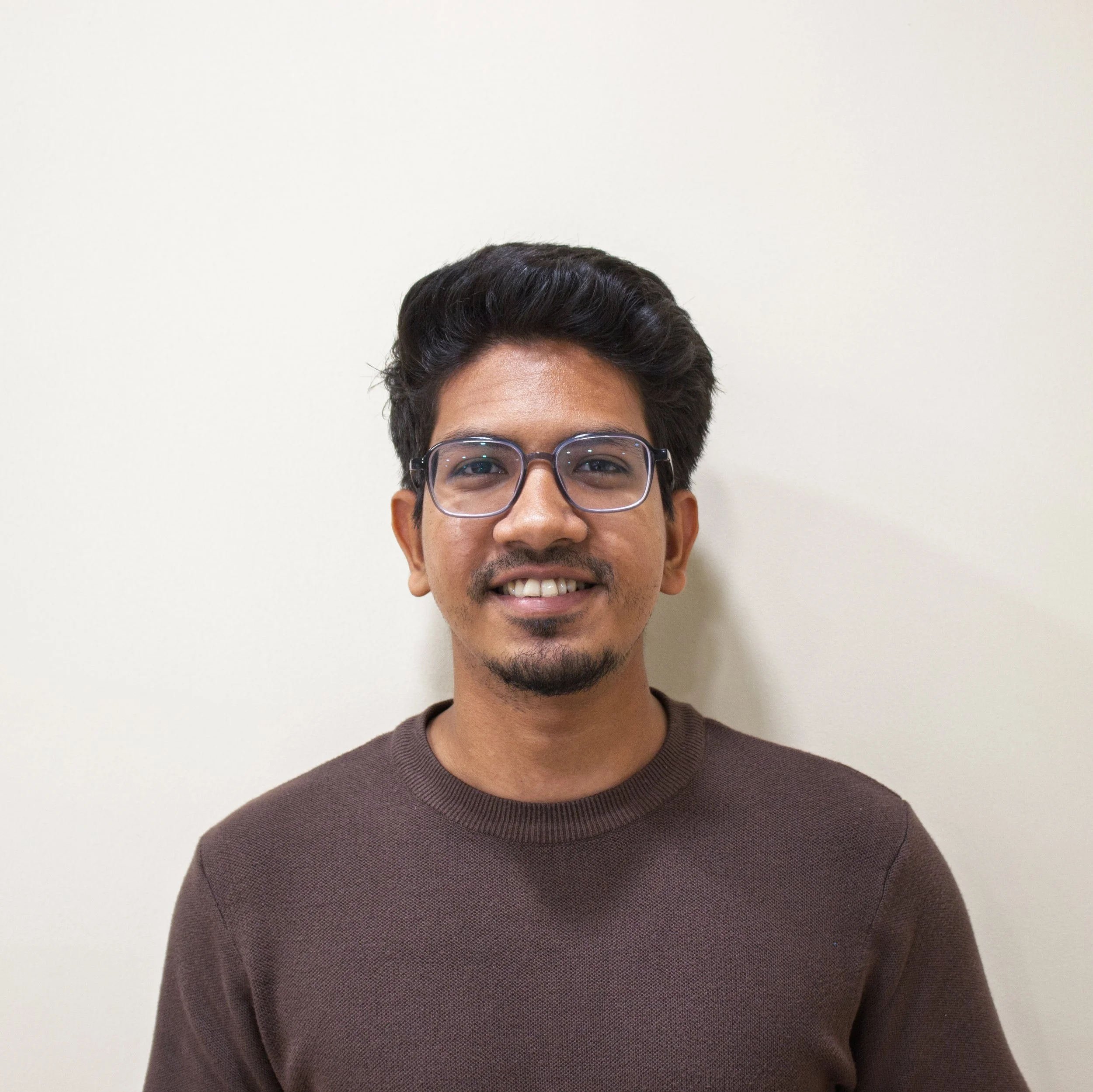 A man with dark hair slicked back, wearing a blue short-sleeve sweater and beige pants, sits on a cream-colored bench with a wooden base, looking to his left with a neutral expression, casting a shadow on the beige wall behind him.