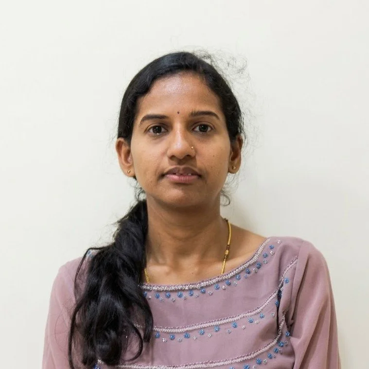 A woman with dark hair in a side braid, wearing a pinkish purple traditional Indian outfit with blue and silver embroidery, standing against a plain white background.