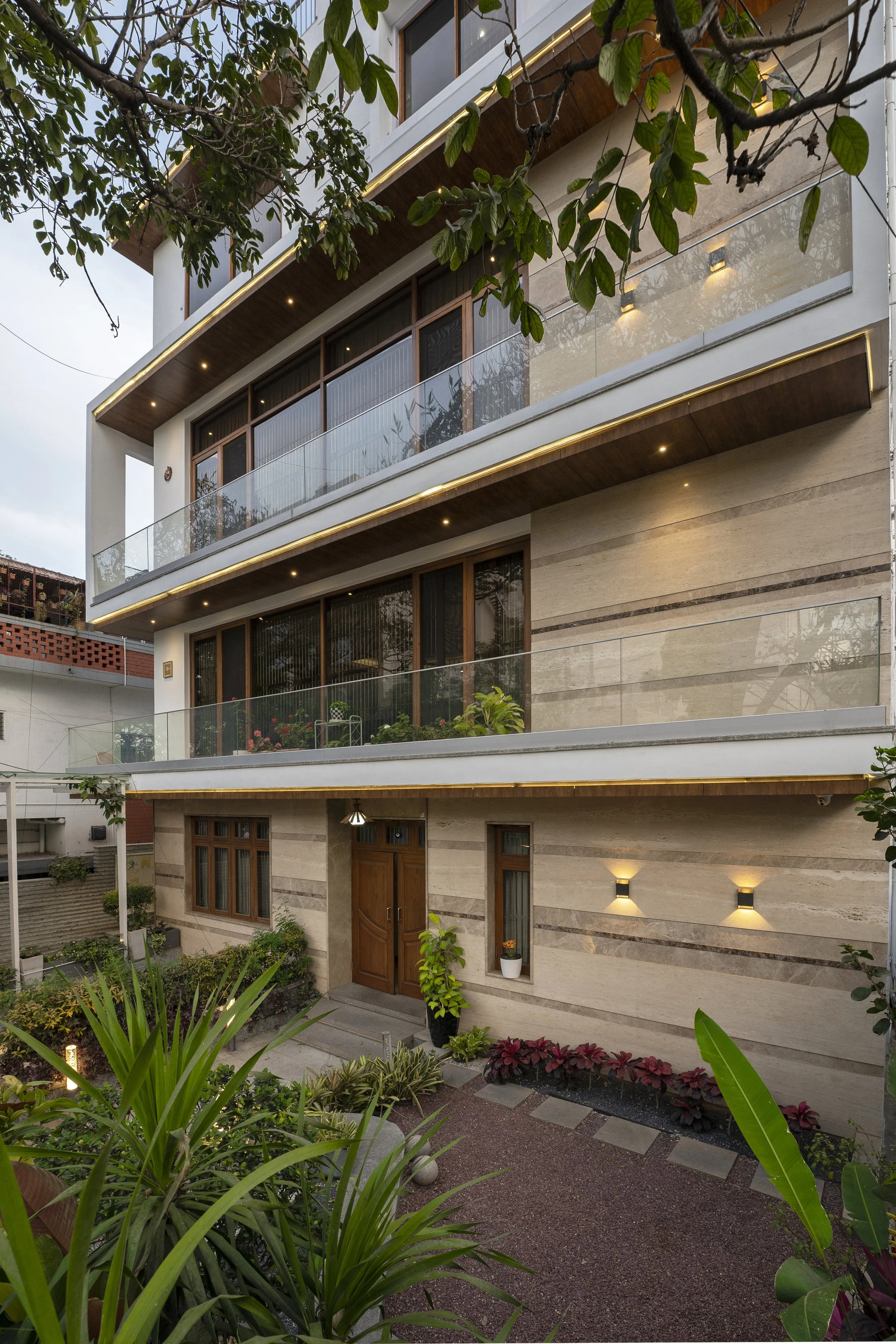 Modern multi-story residential building with large glass windows, balconies with glass railings, warm lighting, and landscaped front yard with plants and stone pathway.