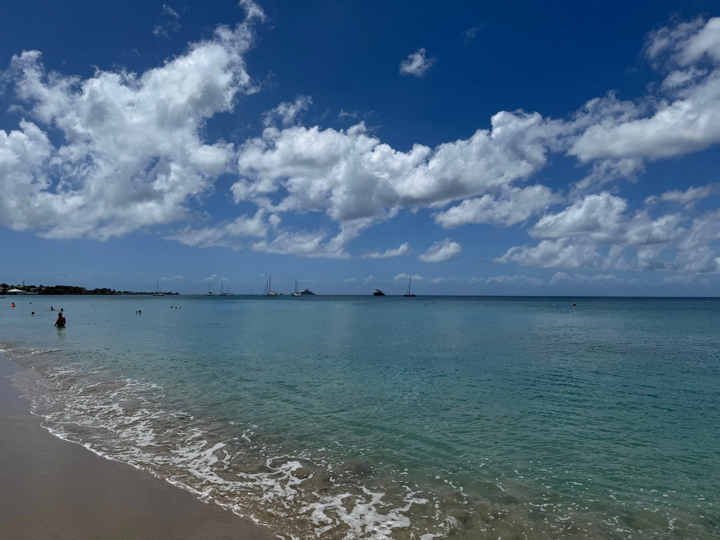 A sunny beach with clear blue water, a few people swimming, and boats anchored in the distance under a sky with scattered clouds.