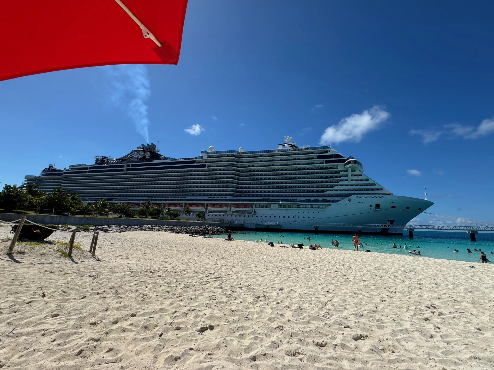 A large cruise ship named MSC Seashore docked near a sandy beach with beachgoers, under a bright blue sky with a few clouds and an orange beach umbrella partially visible in the top left corner.