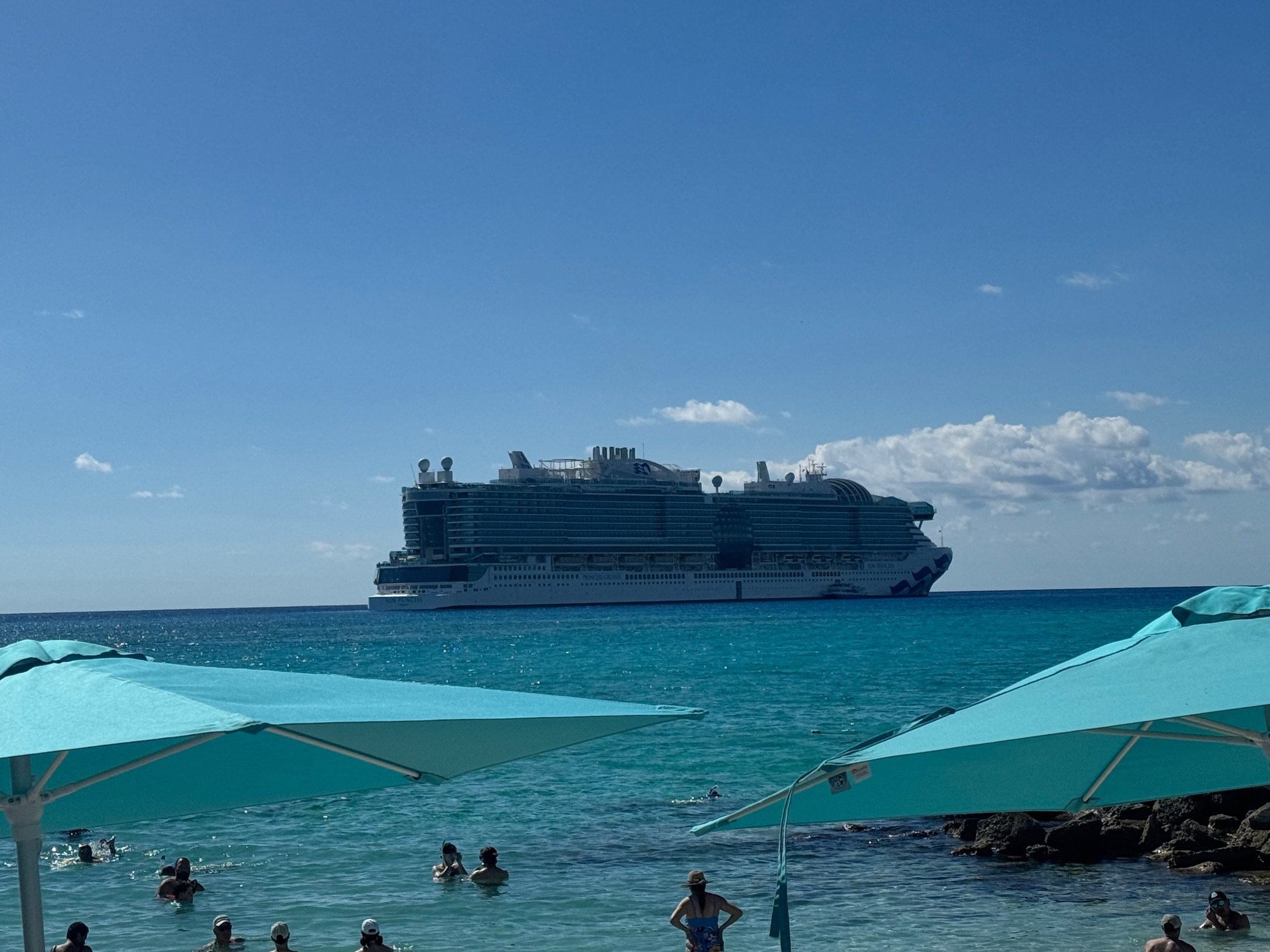 A cruise ship sailing on the ocean viewed from a beach with turquoise umbrellas and people swimming.