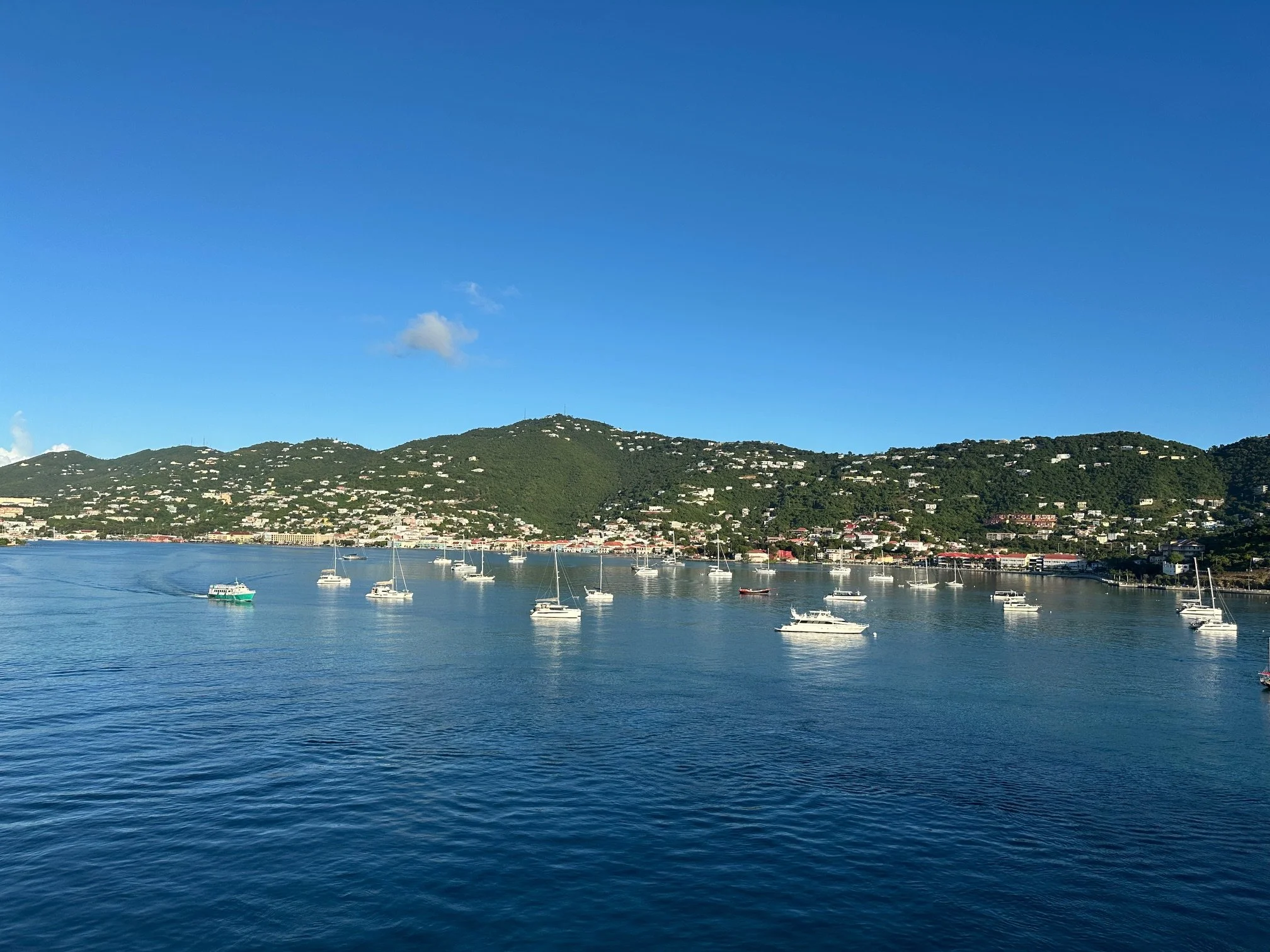A scenic view of a bay with several boats anchored, green hills in the background, and a clear blue sky.