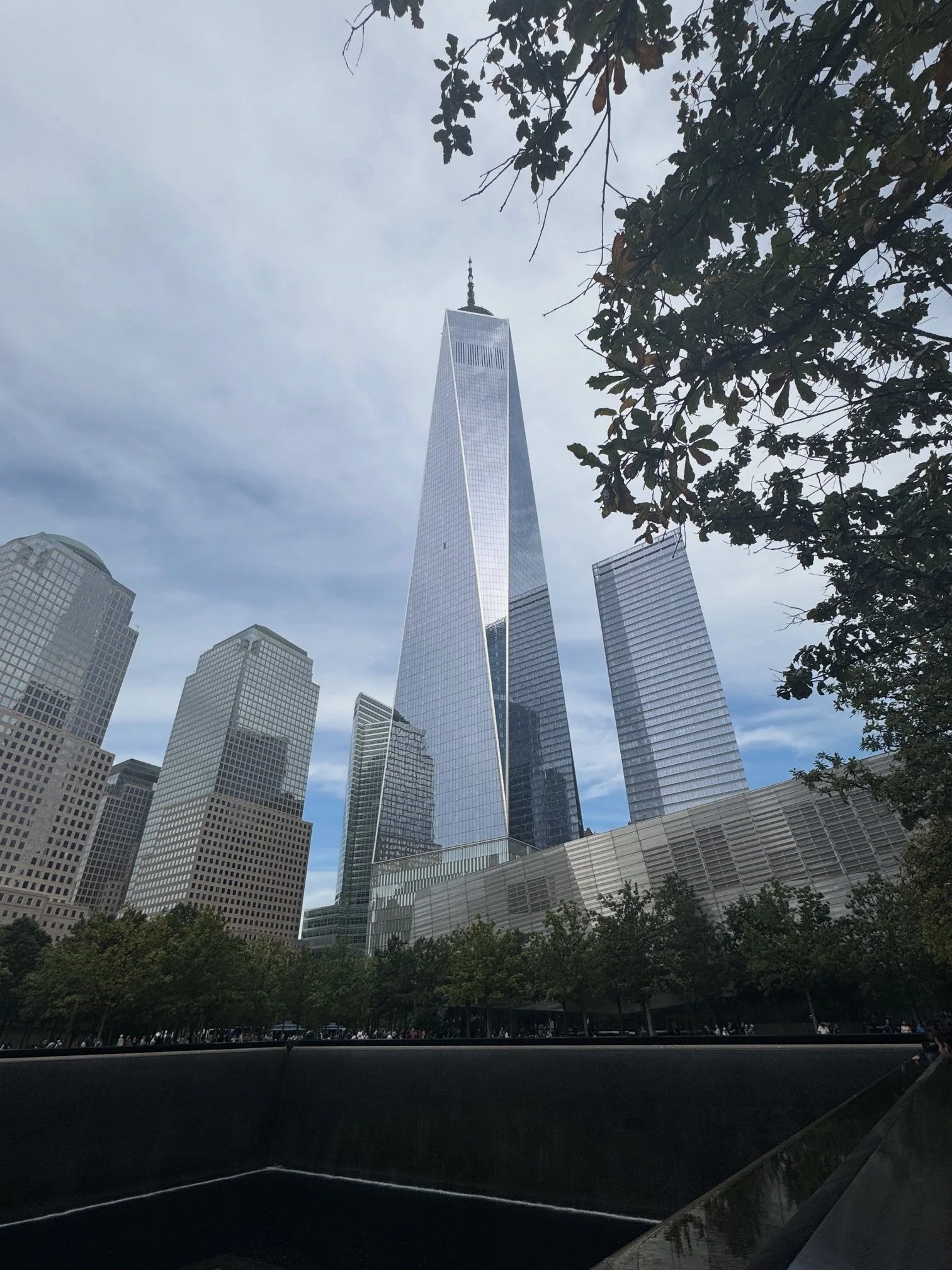 Skyline of Manhattan with One World Trade Center and surrounding skyscrapers with trees in the foreground.