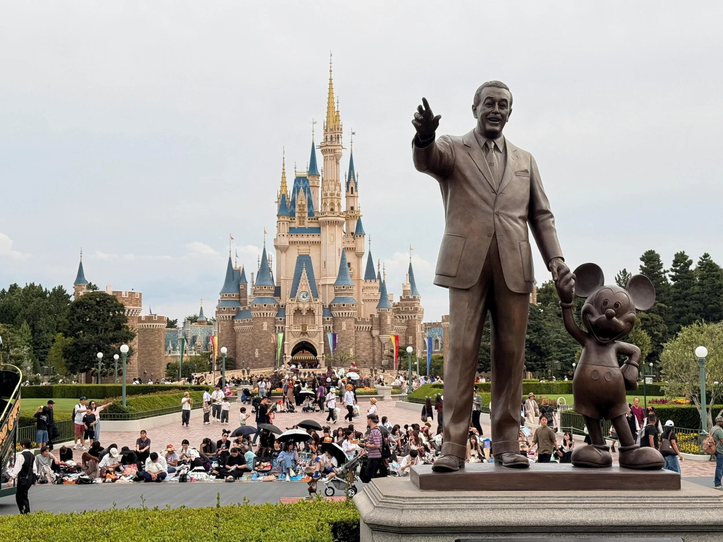 Statue of Walt Disney and Mickey Mouse in front of Sleeping Beauty Castle at Disneyland, with many visitors walking and sitting on the ground.