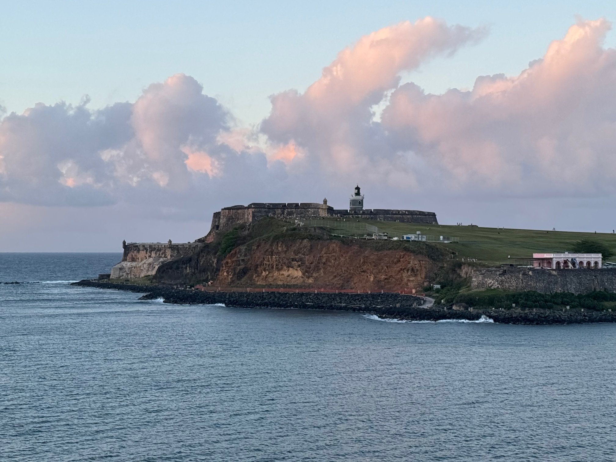 A coastal fortress with a lighthouse on a grassy hill, overlooking the ocean, under a partly cloudy sky during sunset.