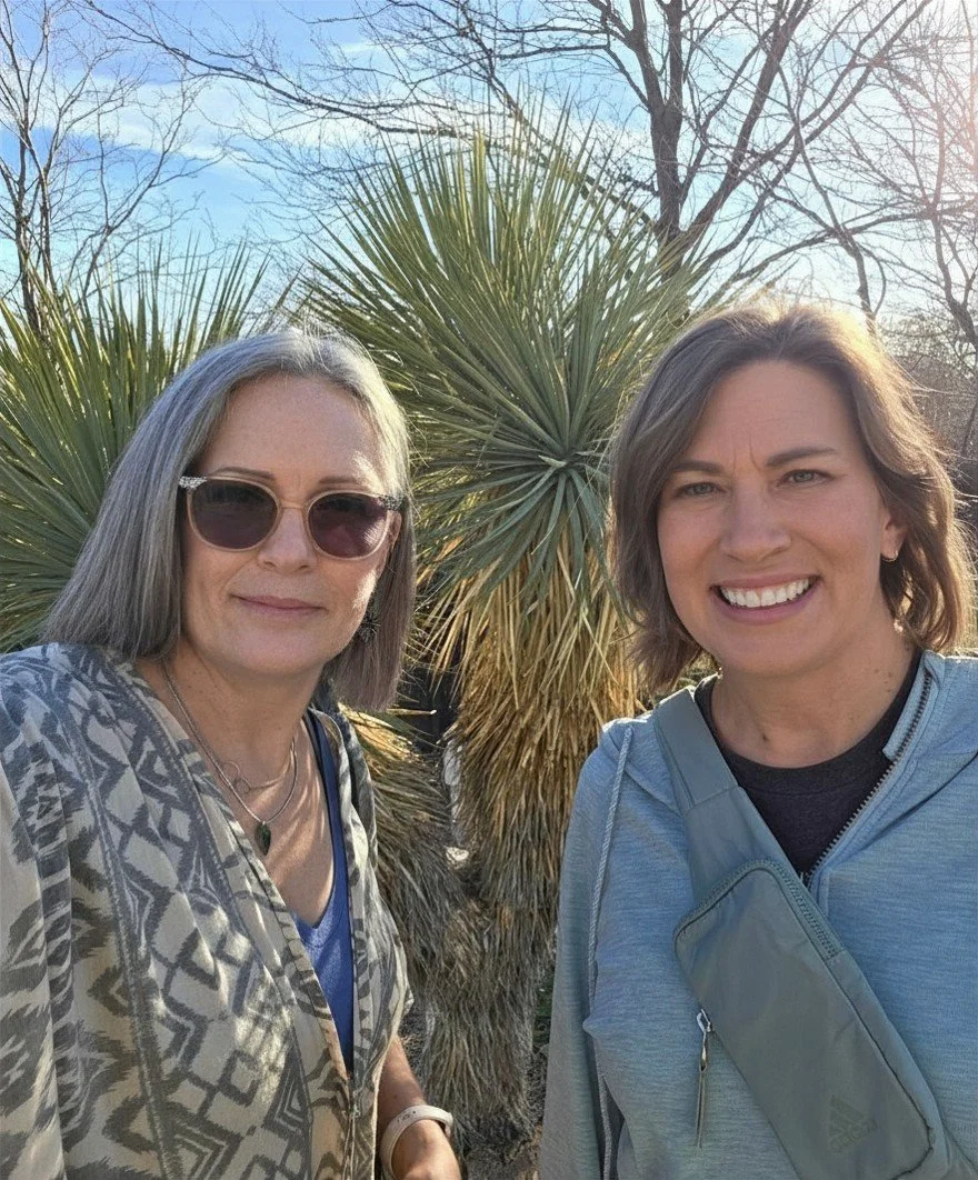 Two women outdoors taking a selfie with a large plant and trees in the background.