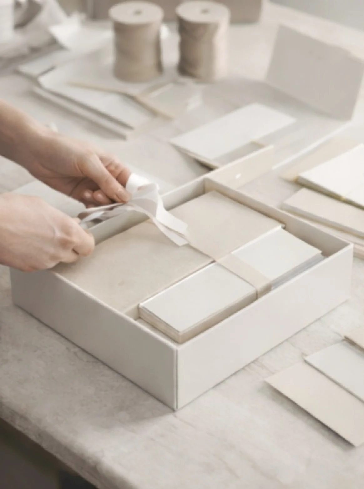 Hands tying a ribbon around a white box of soap bars, with more soap bars and craft materials on a workspace in the background.
