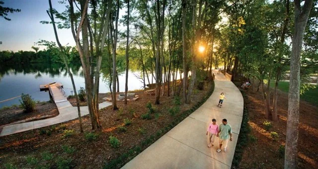 A winding concrete pathway through a wooded park at sunset with people walking and relaxing by a pond.
