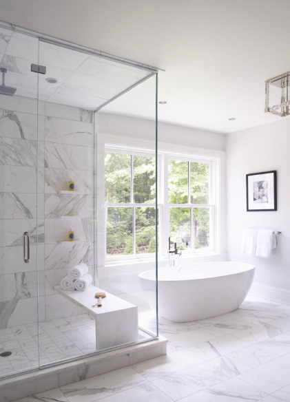 Modern bathroom with glass-enclosed shower, white freestanding bathtub, marble flooring, and large window with views of greenery.