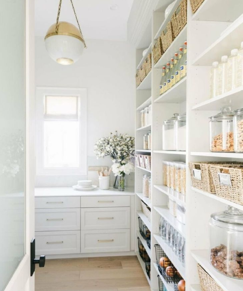 White pantry with organized shelves filled with jars, baskets, and containers of dry goods, and a countertop with a vase of flowers near a window.