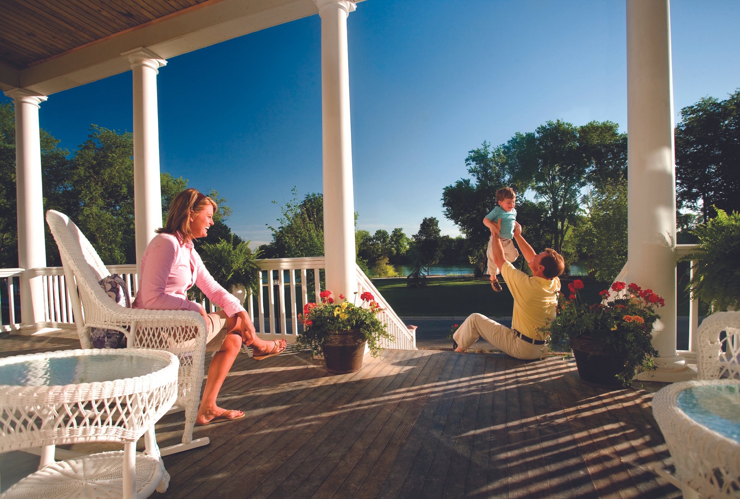A family enjoying a sunny day on a porch with white columns and wicker furniture. A woman is seated, a man is sitting on the floor playing with a young boy who is lifted into the air. Potted plants with flowers decorate the porch, and trees and a body of water are visible in the background.