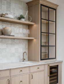 Kitchen with light wooden cabinets, open shelves decorated with vases and plants, a glass-door cabinet, beige countertop, and brass faucet.