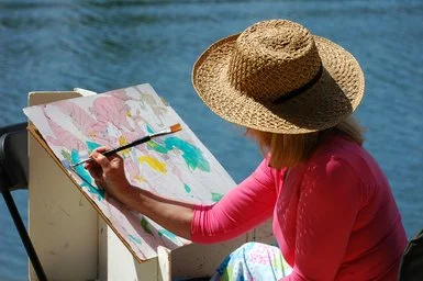 Woman wearing a large straw hat and pink shirt painting with watercolors outdoors near a body of water.