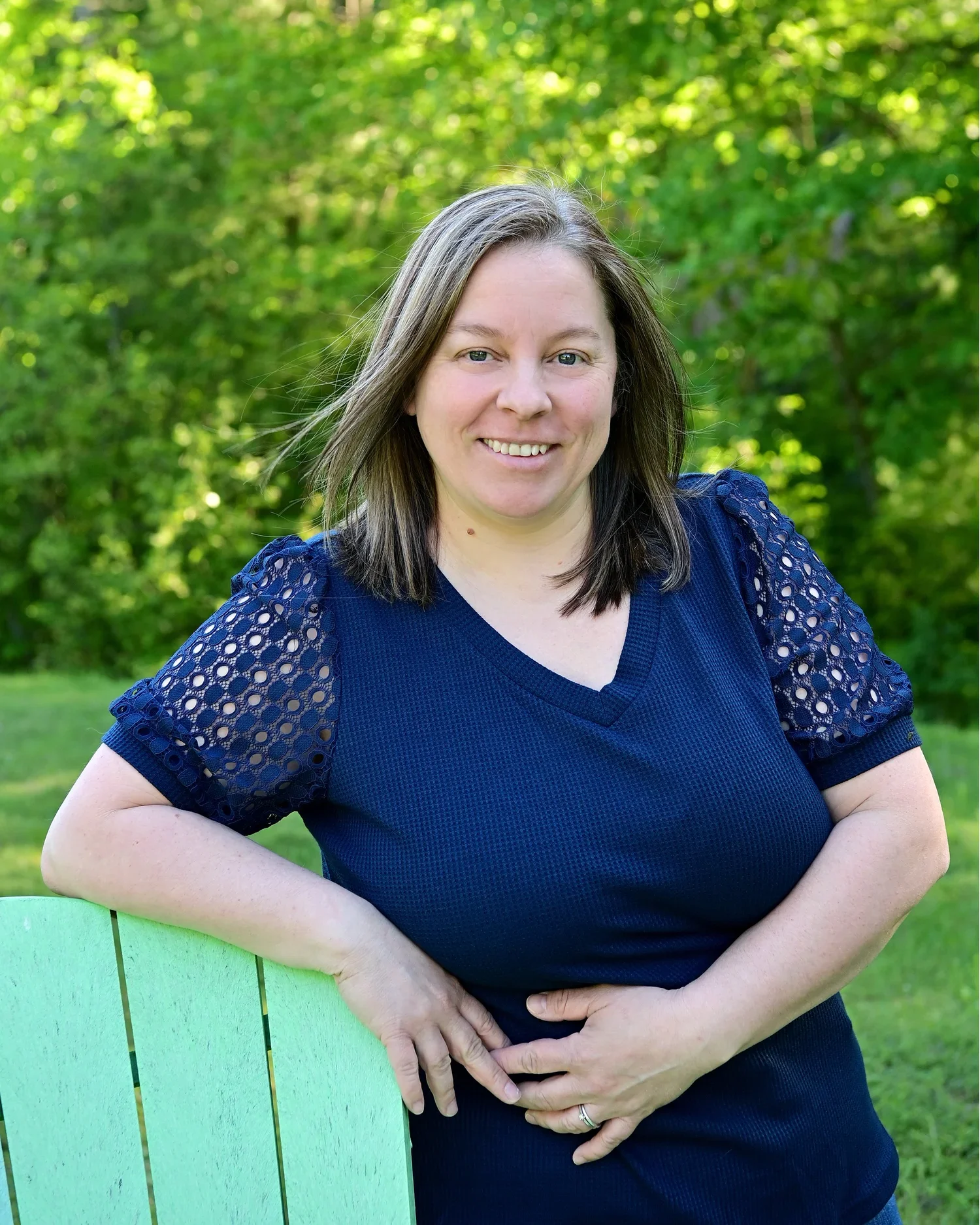 A woman standing outdoors near a green painted bench, wearing a navy blue top with lace sleeves, smiling, with trees and greenery in the background.