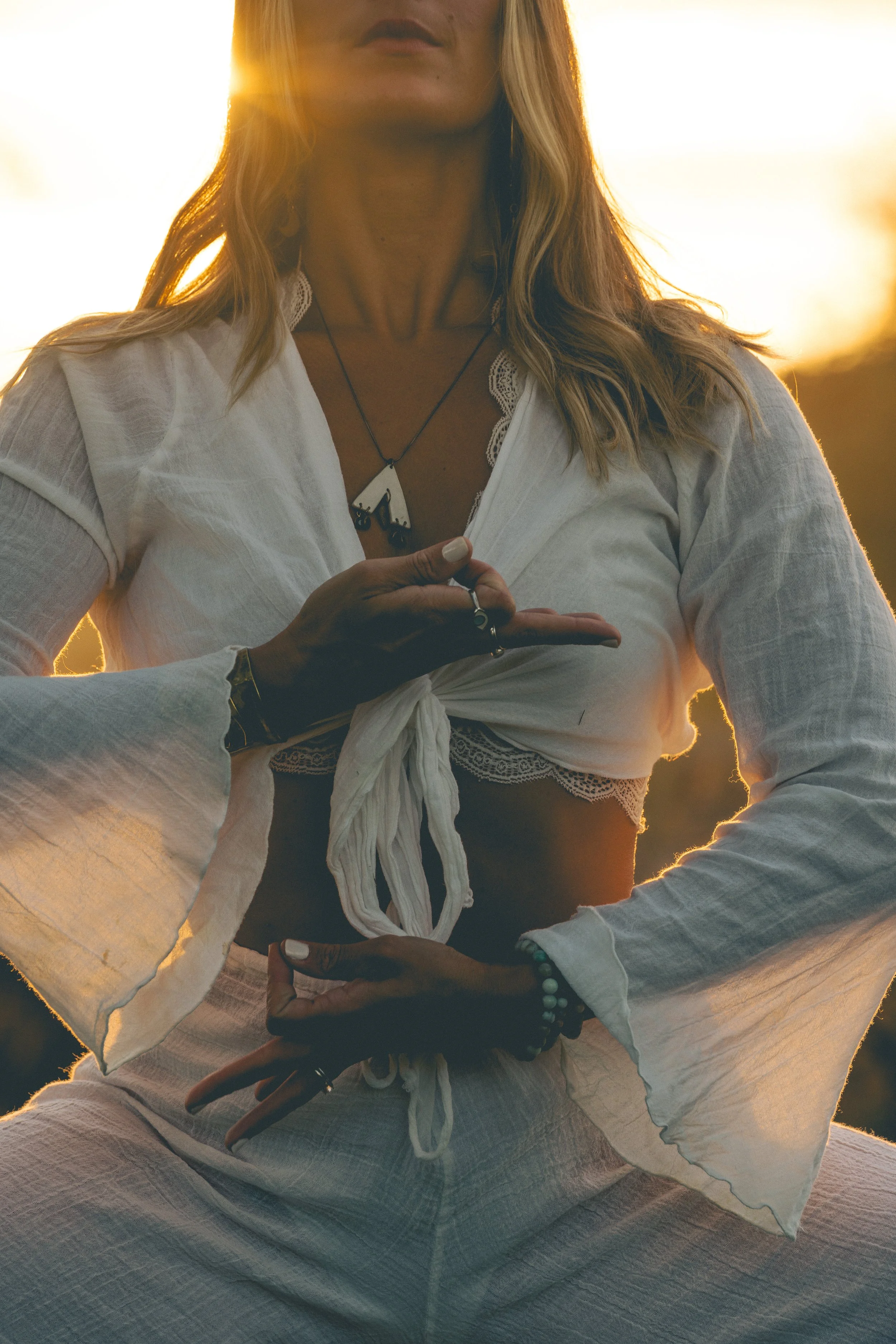 A woman with long blonde hair is sitting cross-legged during sunset, wearing a white top and beige pants, with her hands resting on her lap and her gaze directed forward.