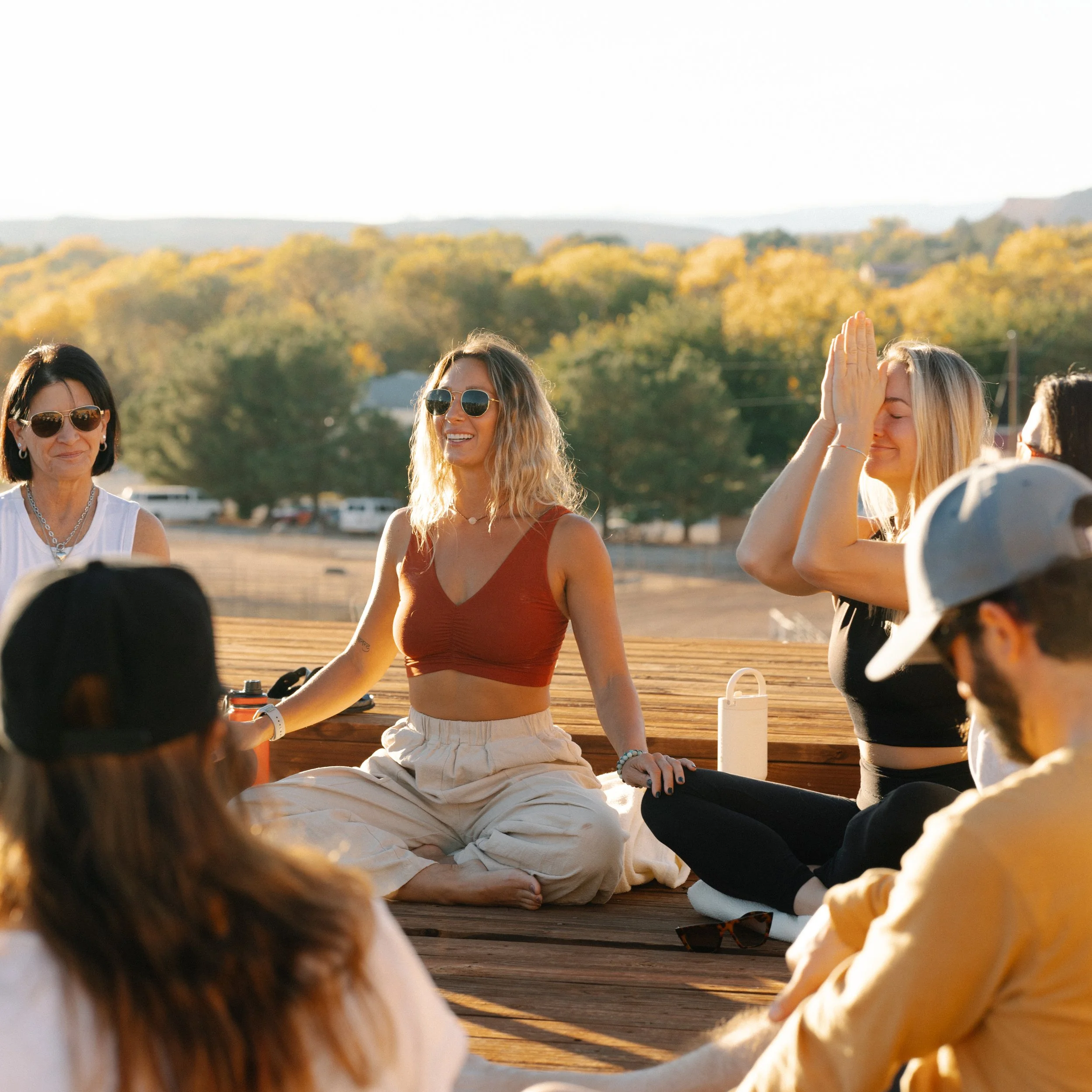 A group of women sitting outdoors on a wooden platform, participating in a meditation or yoga session, with trees and hills in the background during sunset.