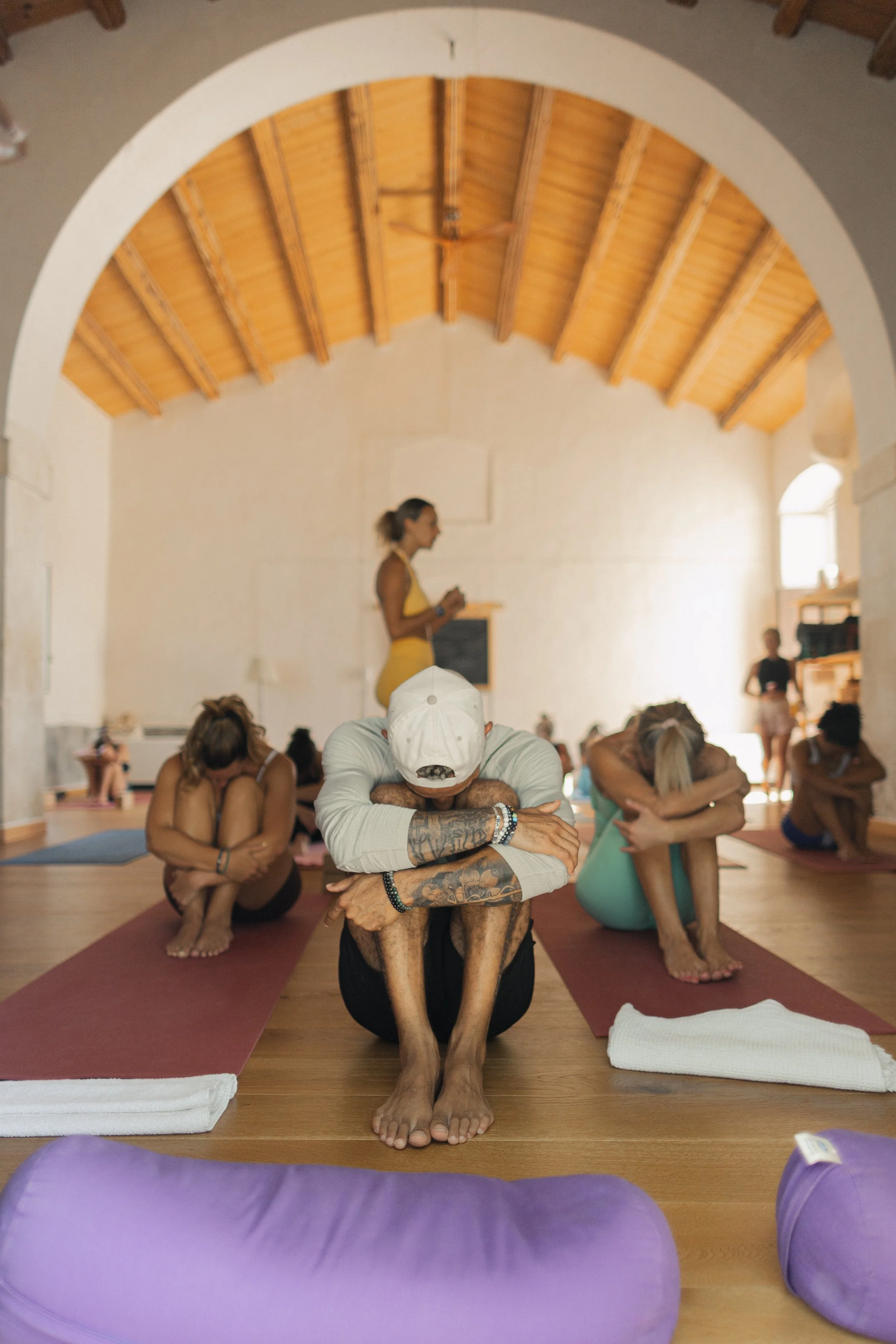 People participating in a yoga or meditation class in a spacious room with wooden ceiling beams. They are sitting cross-legged on yoga mats, with some using their arms to stretch or relax. The room has natural light coming in from windows.