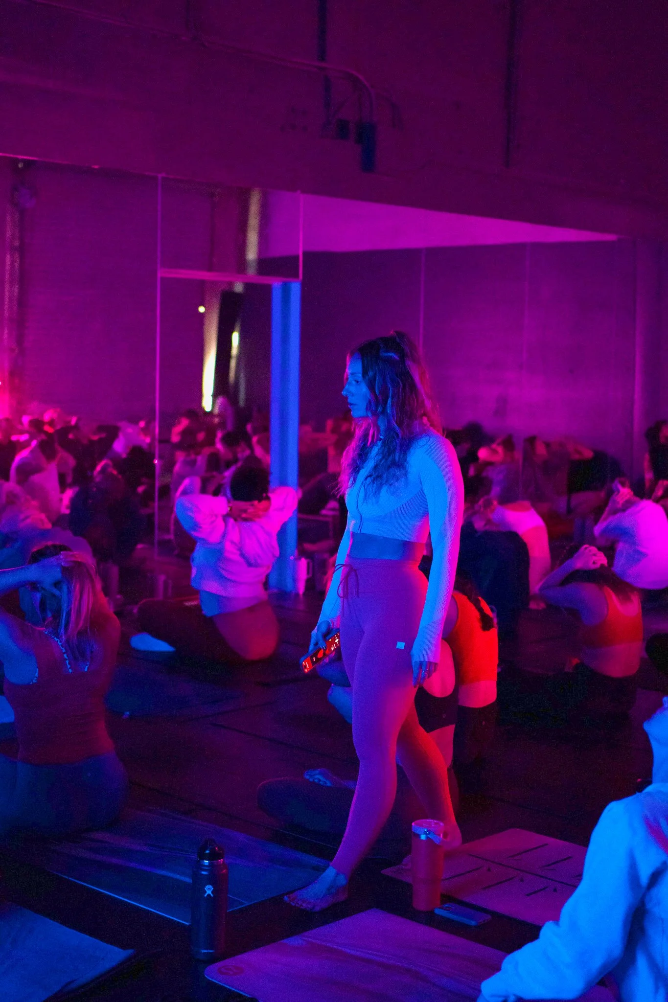 A woman practicing yoga in a dimly lit room with purple and pink lighting. She is standing on a yoga mat, holding a yoga pose, surrounded by other people seated on yoga mats, possibly participating in a class or event.
