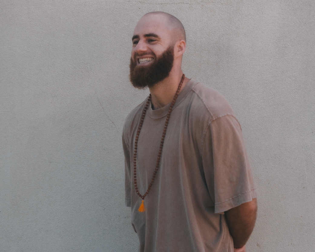 A smiling man with a beard and shaved head standing against a plain wall, wearing a casual t-shirt and a beaded necklace.