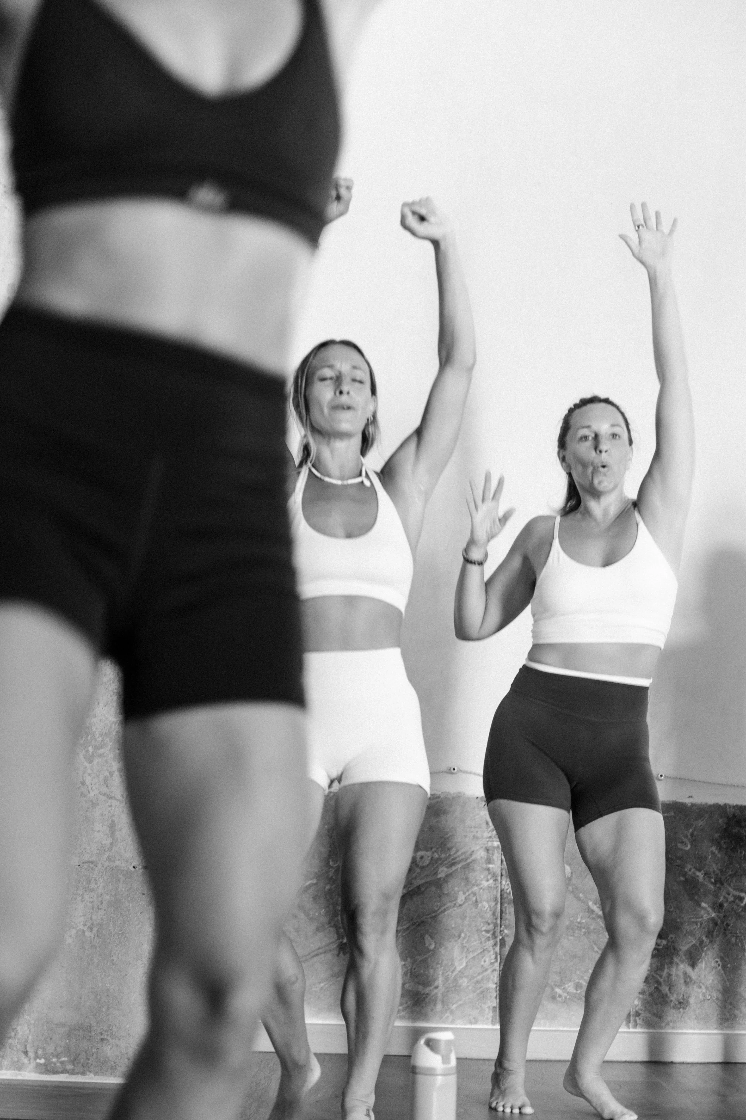 Black and white photo of three women in athletic wear, standing and stretching against a wall with a concrete texture, with one woman in the foreground partially out of focus.