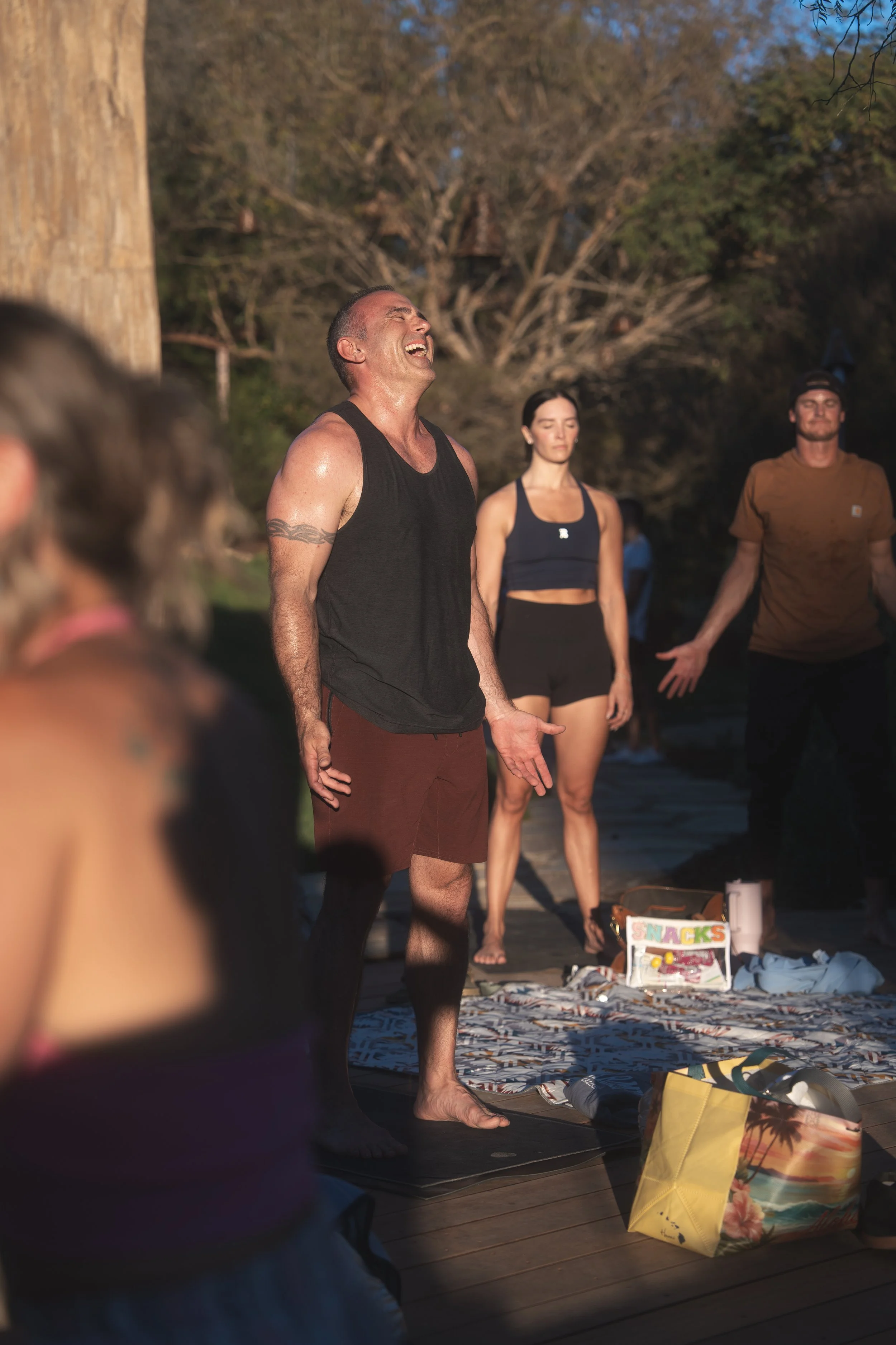 A group of people practicing yoga outdoors on a sunny day, with a man in a black tank top standing with his eyes closed and arms outstretched, surrounded by others, some with their eyes closed. There are trees and a snack box on the ground.