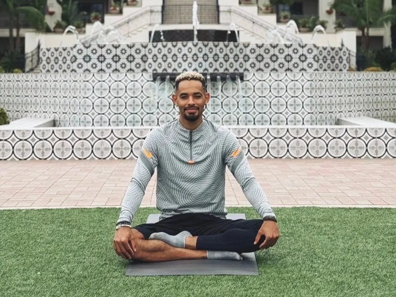 A man practicing yoga outdoors on a black mat with a patterned tiled staircase in the background.