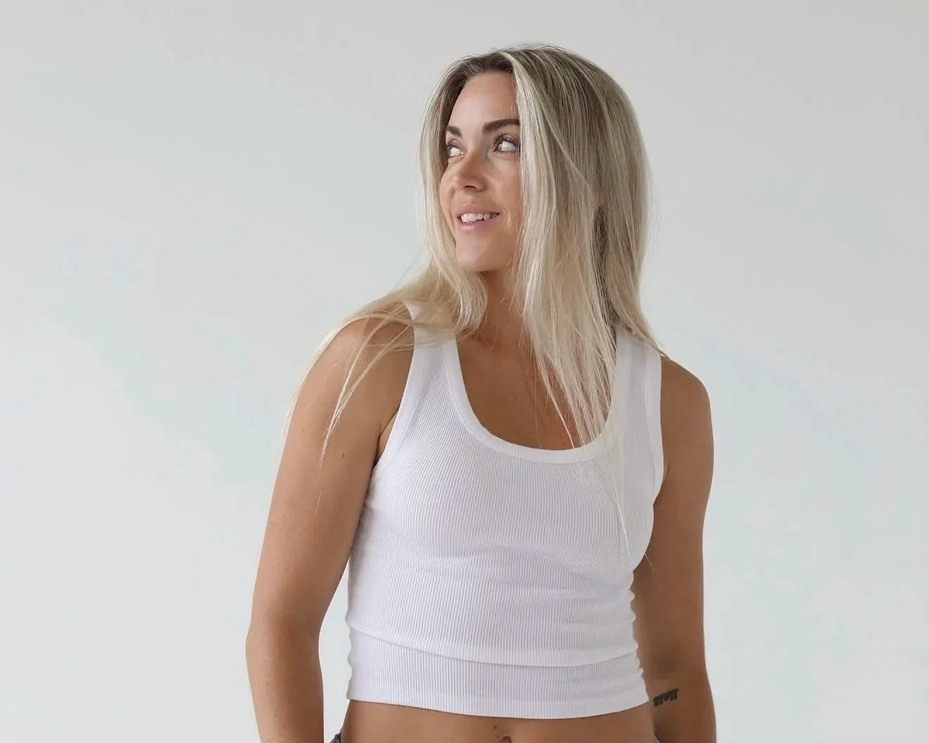 A woman with blonde, shoulder-length hair wearing a white tank top, looking to her left with a slight smile, standing against a plain white background.
