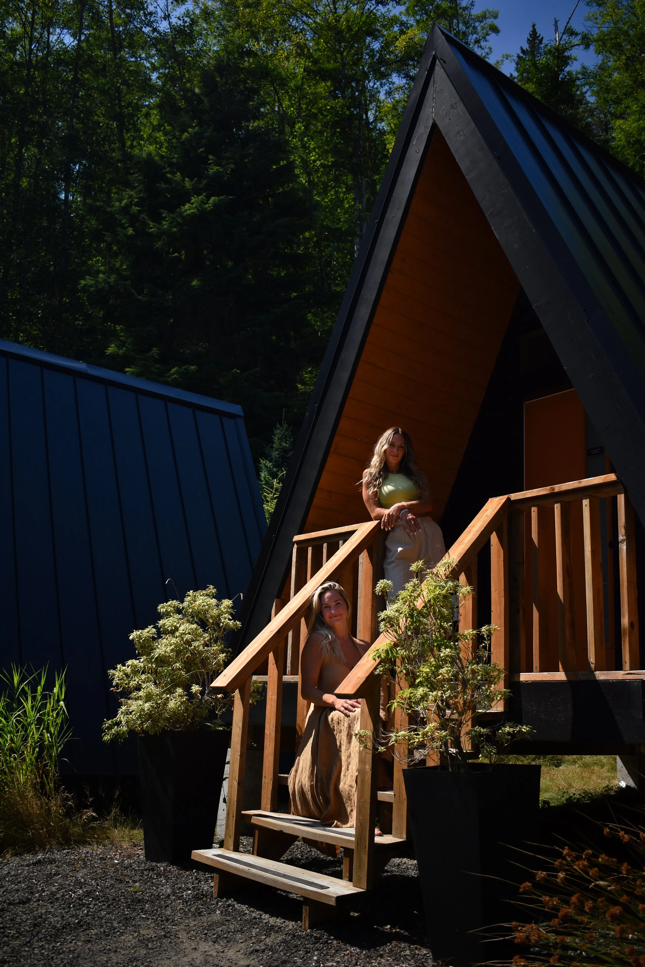 Two women standing on wooden stairs in front of A-frame cabins surrounded by greenery and trees.