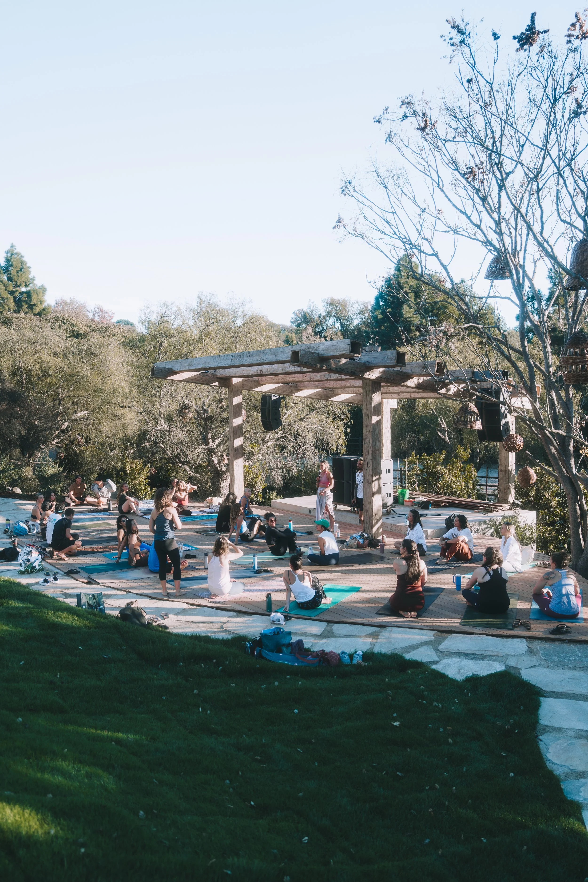 A group of people participating in an outdoor yoga class on yoga mats in a park during daylight, with trees and a wooden canopy structure in the background.