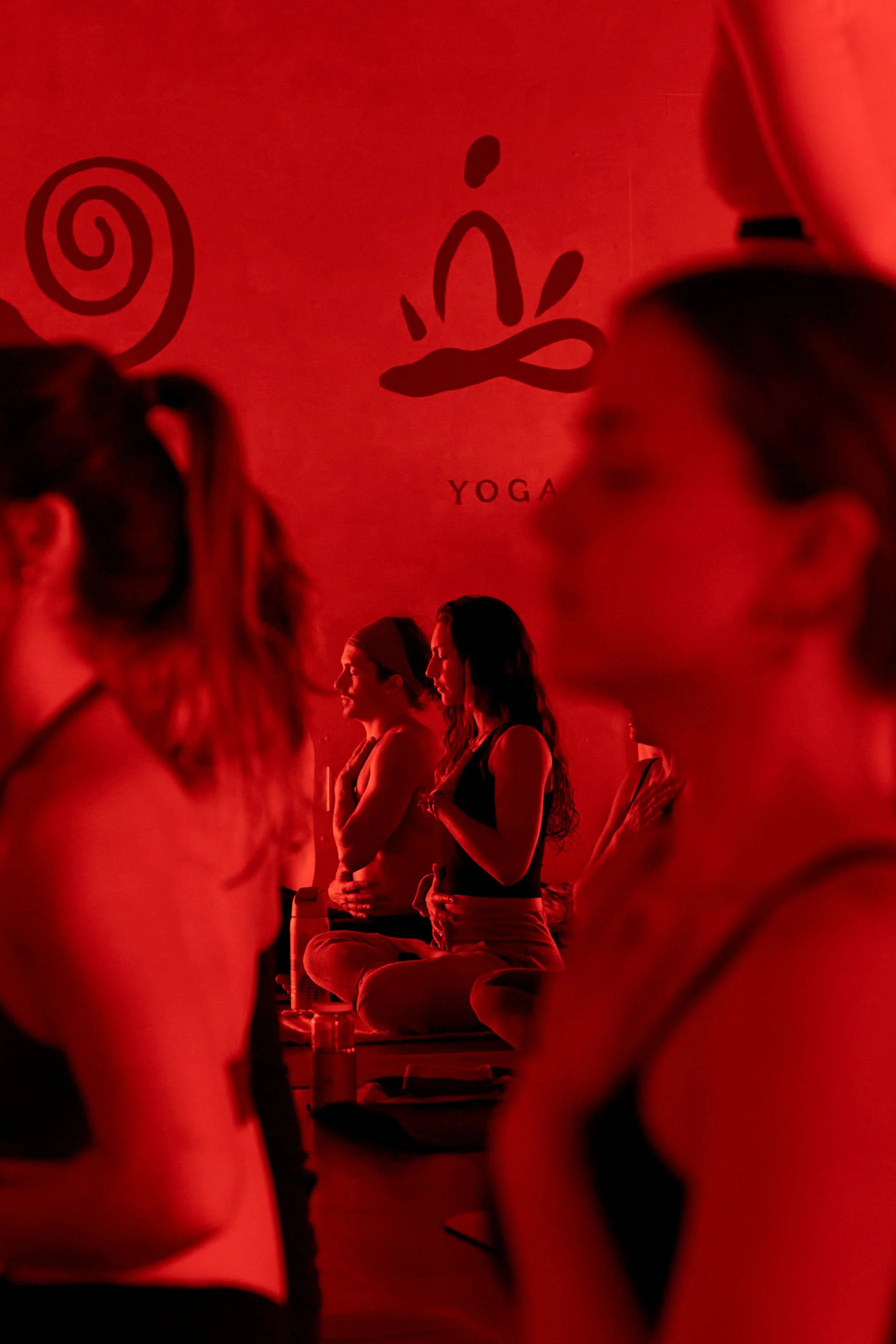People participating in a yoga class in a dimly lit room with red lighting, with a sign reading 'YOGA' on the wall in the background.