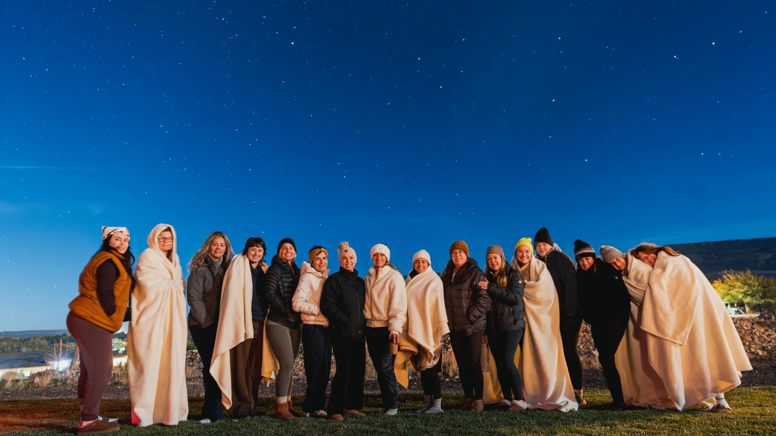 Group of people standing outdoors at night under a starry sky, some of them wrapped in blankets and wearing hats and jackets.