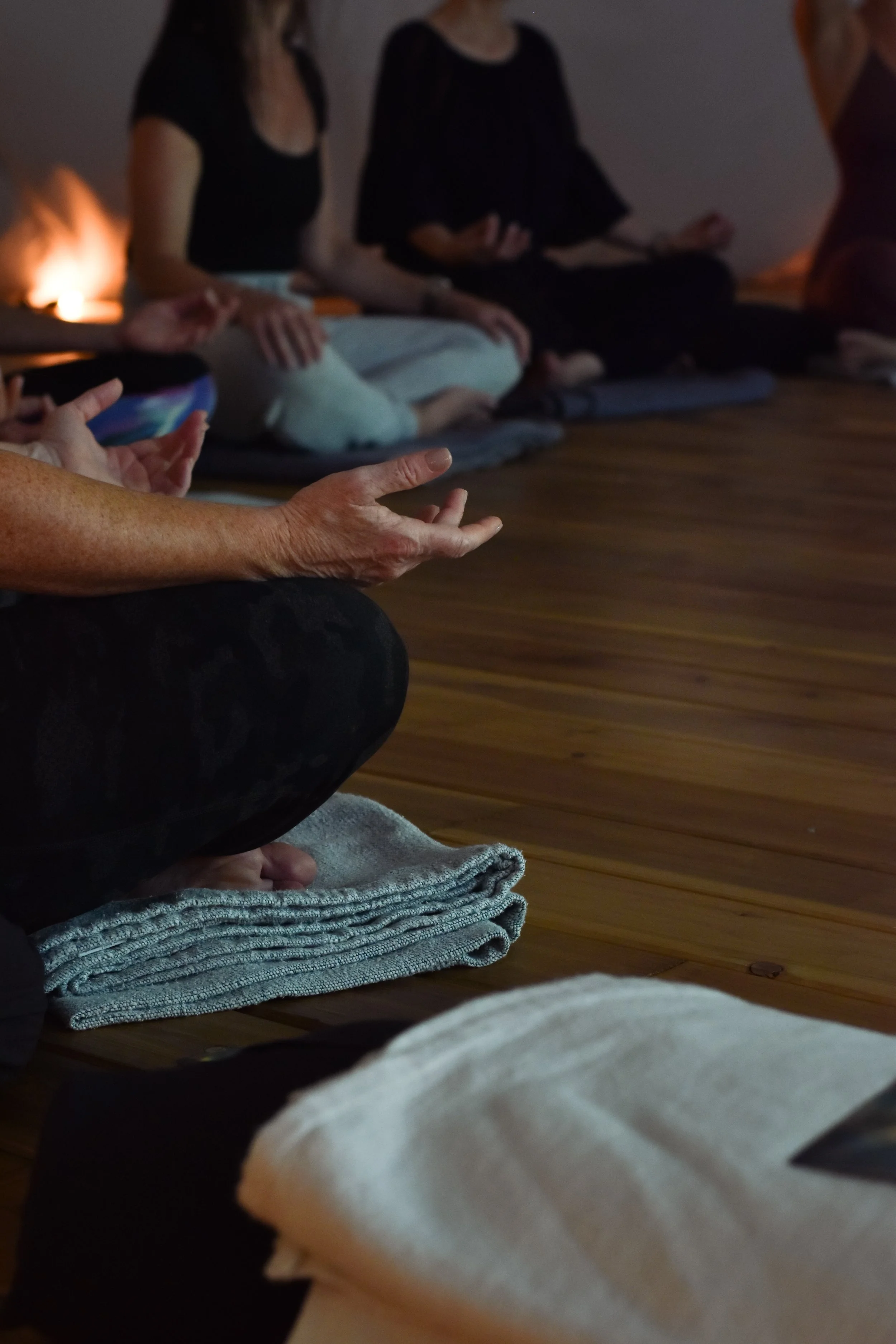 People sitting in a circle in a dimly lit room, practicing meditation, with a wooden floor and blankets underneath.